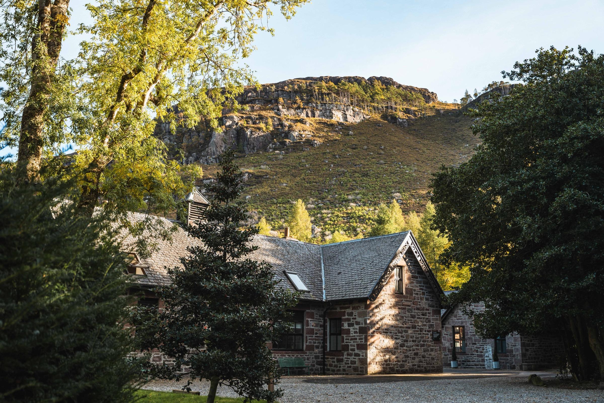 A stone outbuilding with a slate roof among trees, set below a rocky, tree lined hillside.