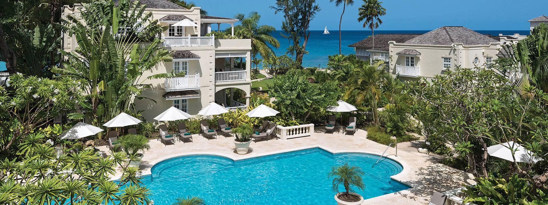 An aerial view of the hotel's cream buildings and balconies among palm trees, with a curved pool, white loungers and umbrellas, and the sea and a sailboat beyond.