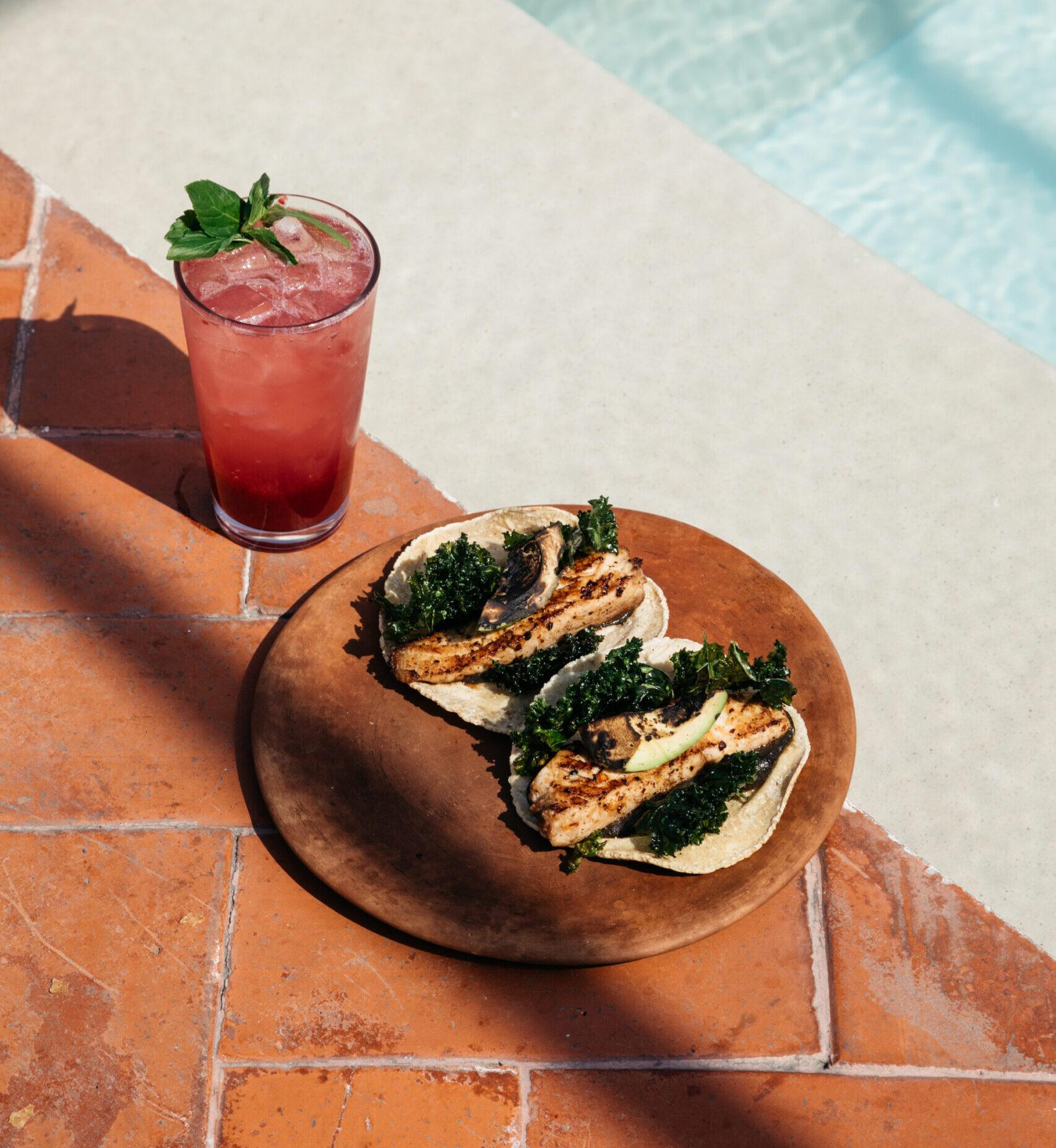 Two fish tacos on a wooden plate beside a pink iced drink with a mint sprig, set on terracotta tiles next to a pool edge.
