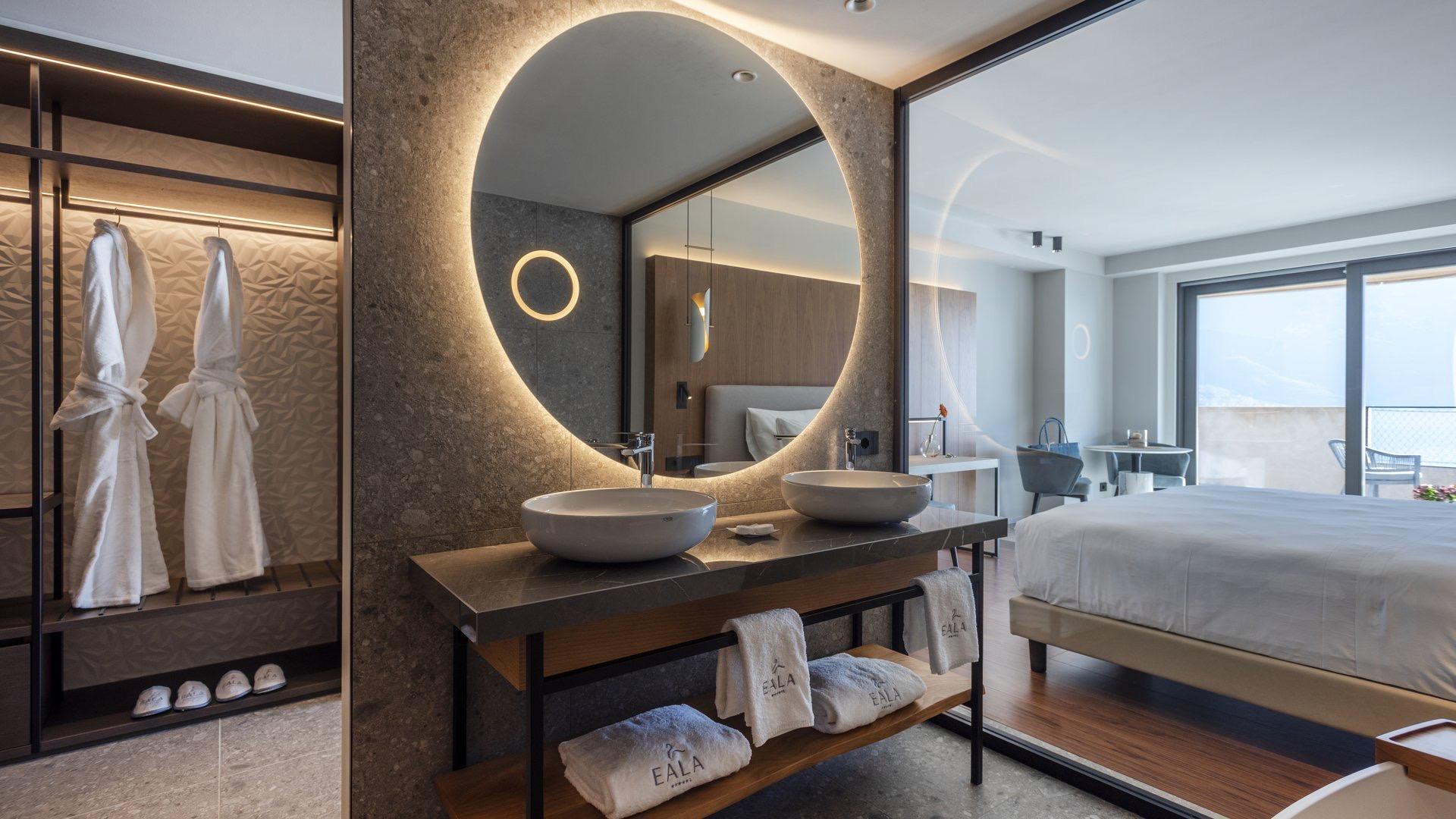 a hotel bathroom with a round backlit mirror and twin basins, opening onto a bedroom with a made bed and a view beyond