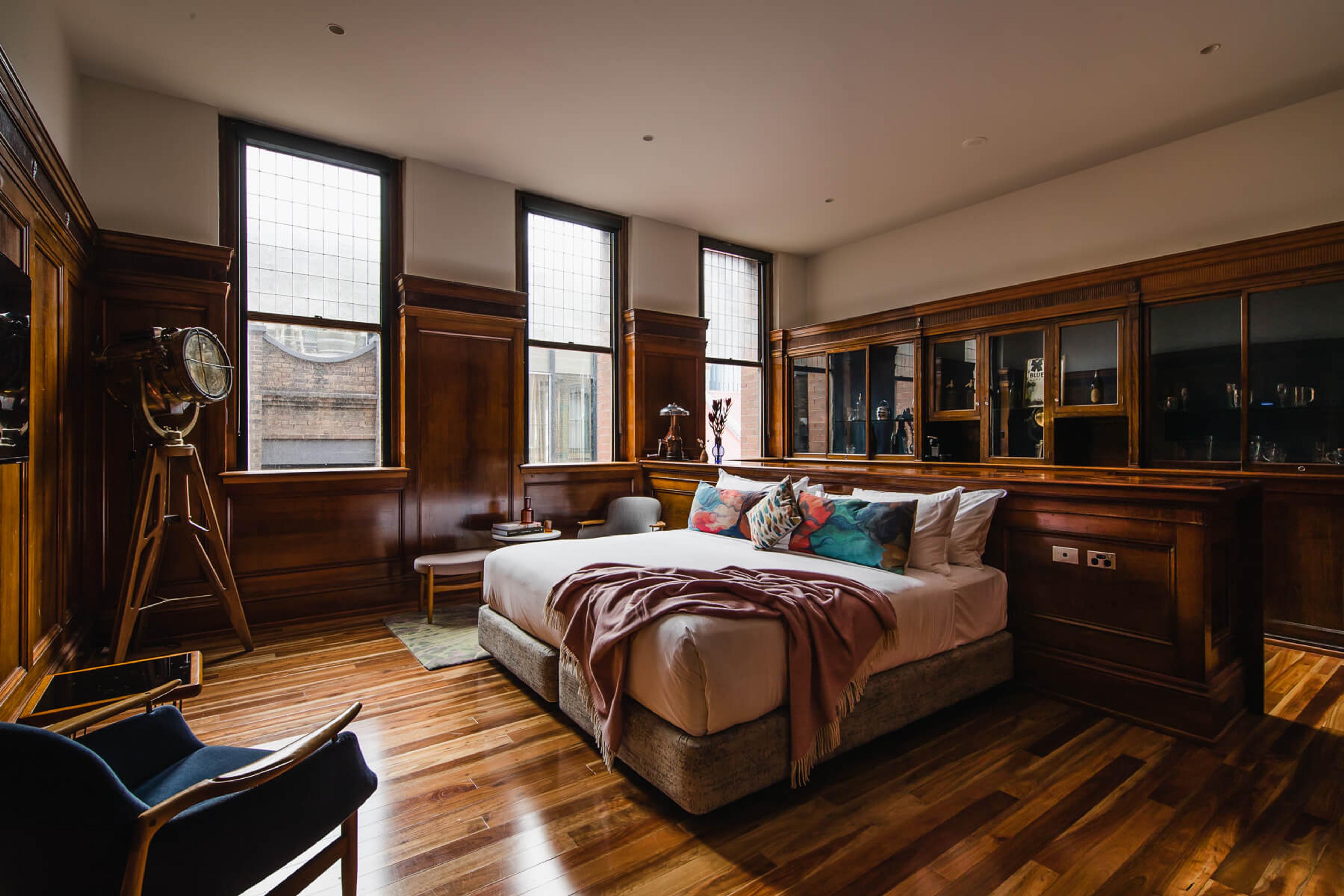 A wood-panelled bedroom with three leadlight windows, a bed with colourful cushions and a blush throw, a brass spotlight lamp, and a navy armchair.