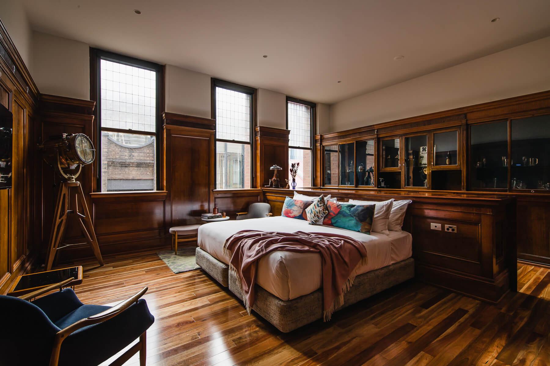 A wood-panelled bedroom with three leadlight windows, a bed with colourful cushions and a blush throw, a brass spotlight lamp, and a navy armchair.