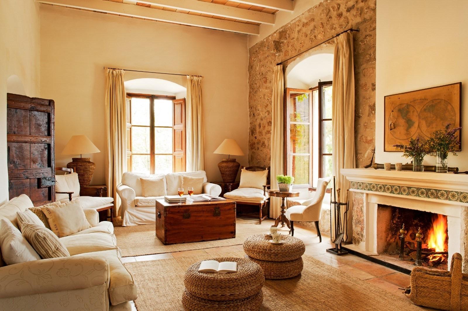 A high-beamed stone living room with white sofas, a lit fireplace, a world map above the mantel, and woven stools in front.