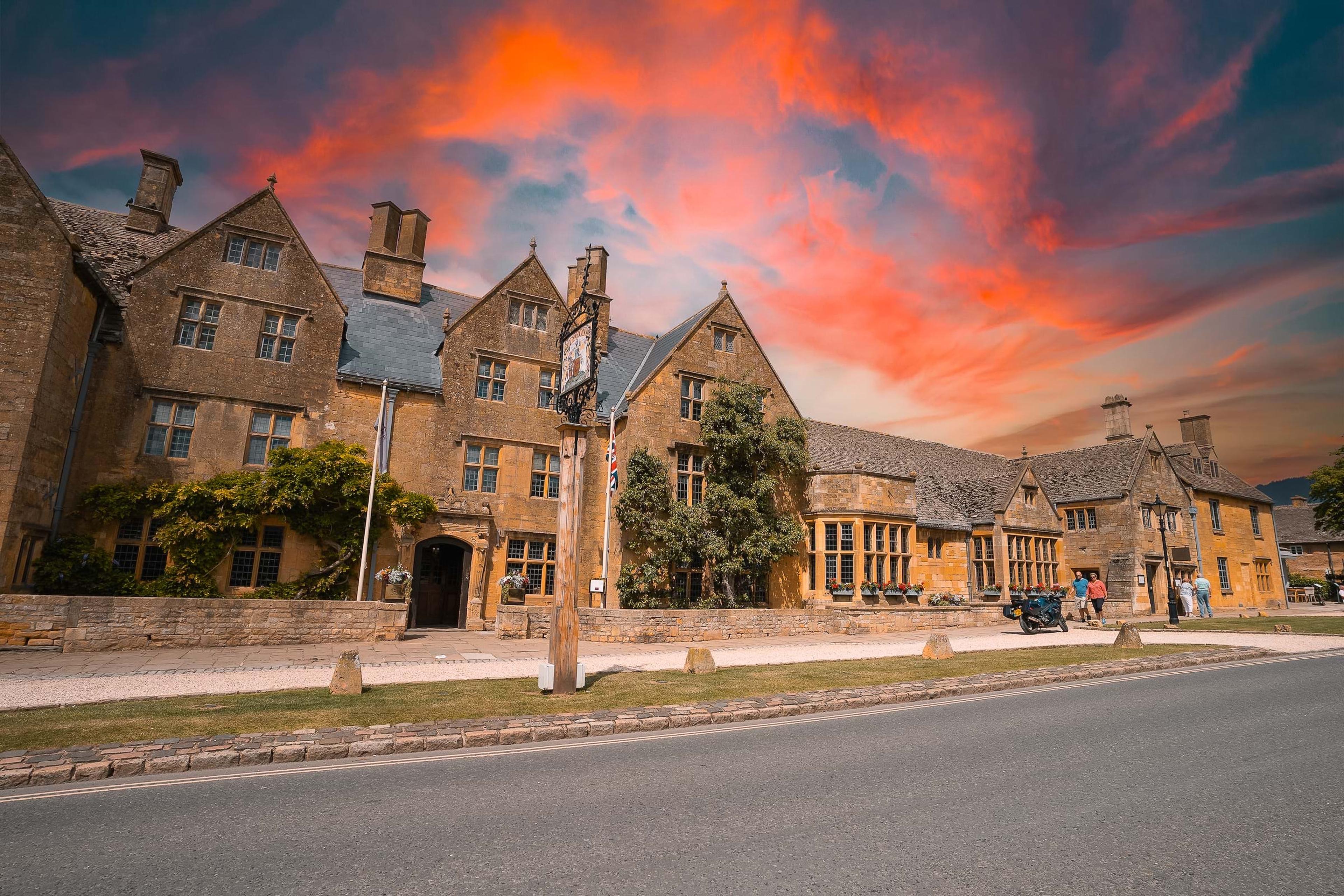 The hotel's stone facade and gabled roofline along the high street, under a vivid orange and pink evening sky, with a few people and a parked motorbike at the roadside.