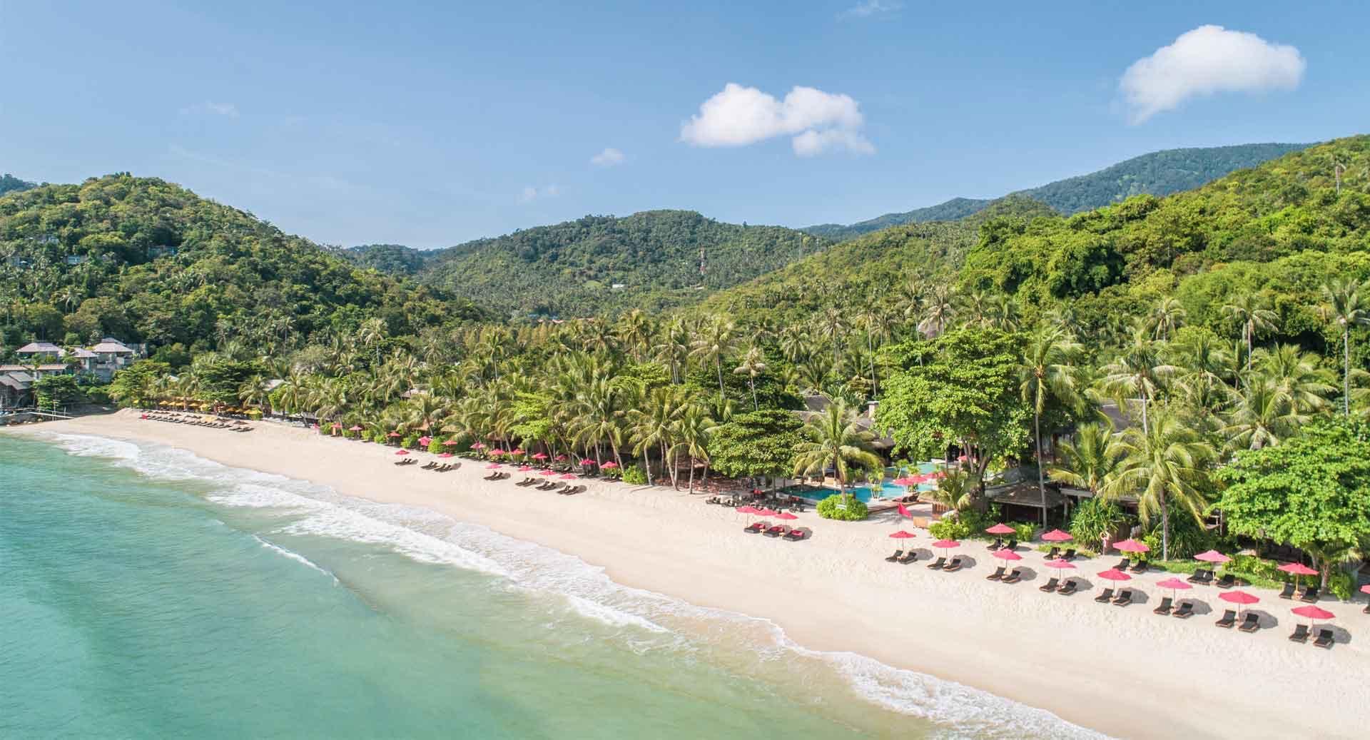 A palm-lined beach and resort buildings seen from above, set against a curving bay and forested hills.