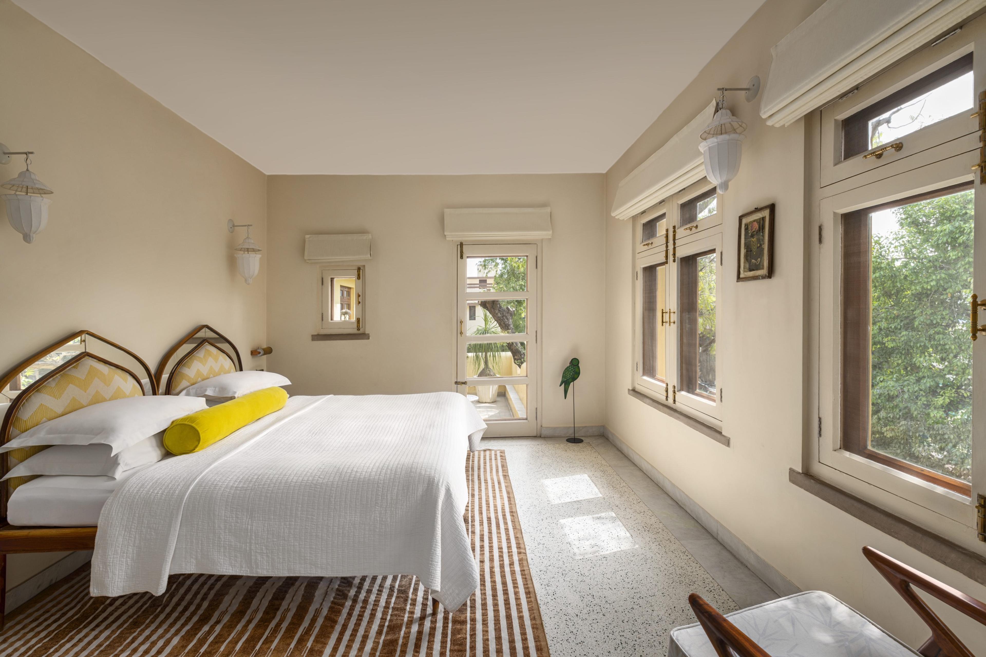 A bright bedroom with a striped rug, a yellow bolster on the bed and French doors open to a terrace with trees beyond.