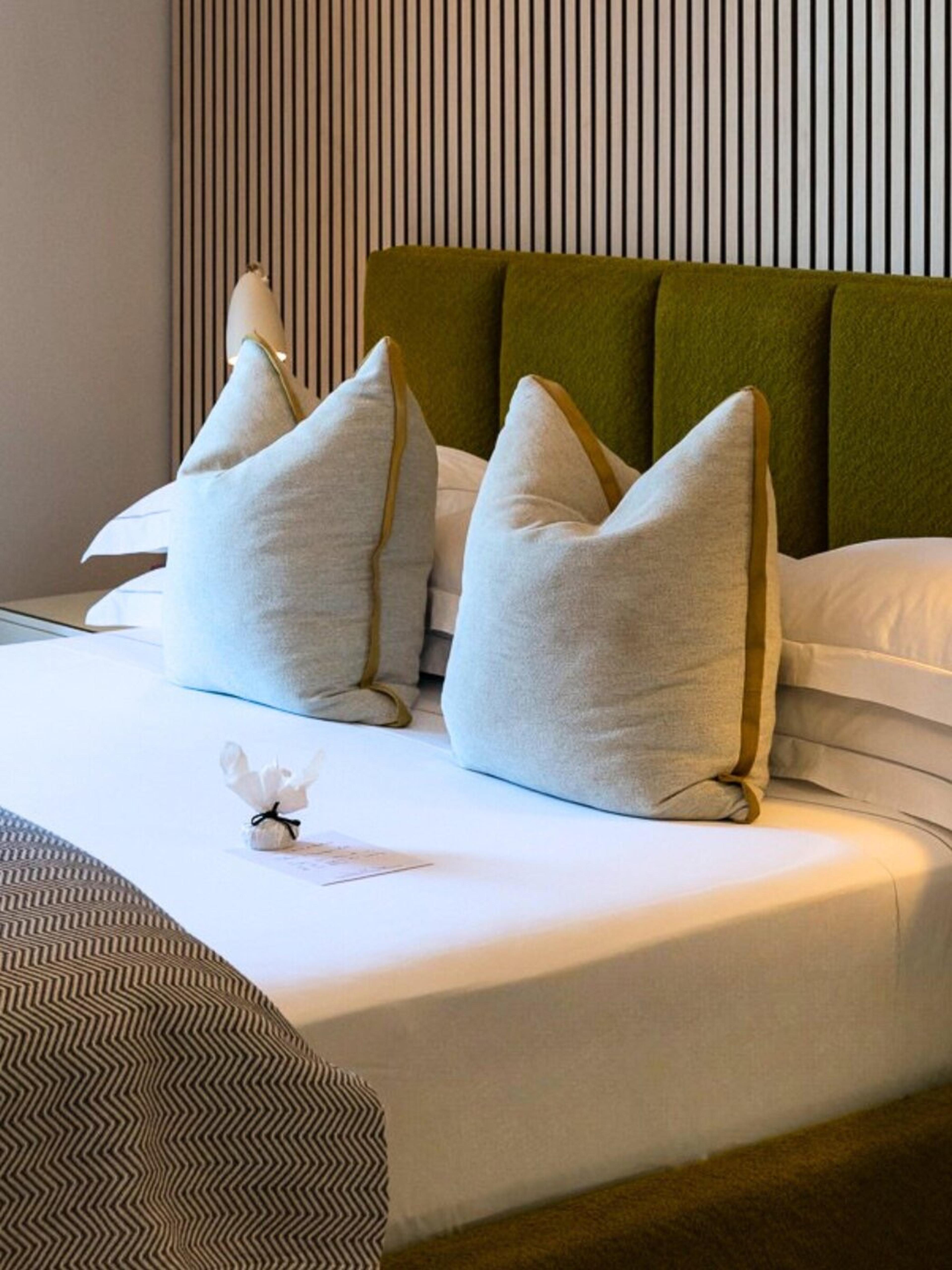 A bed with a green upholstered headboard and two grey cushions on white linen, with a small wrapped sweet on the sheet and a slatted timber wall behind.