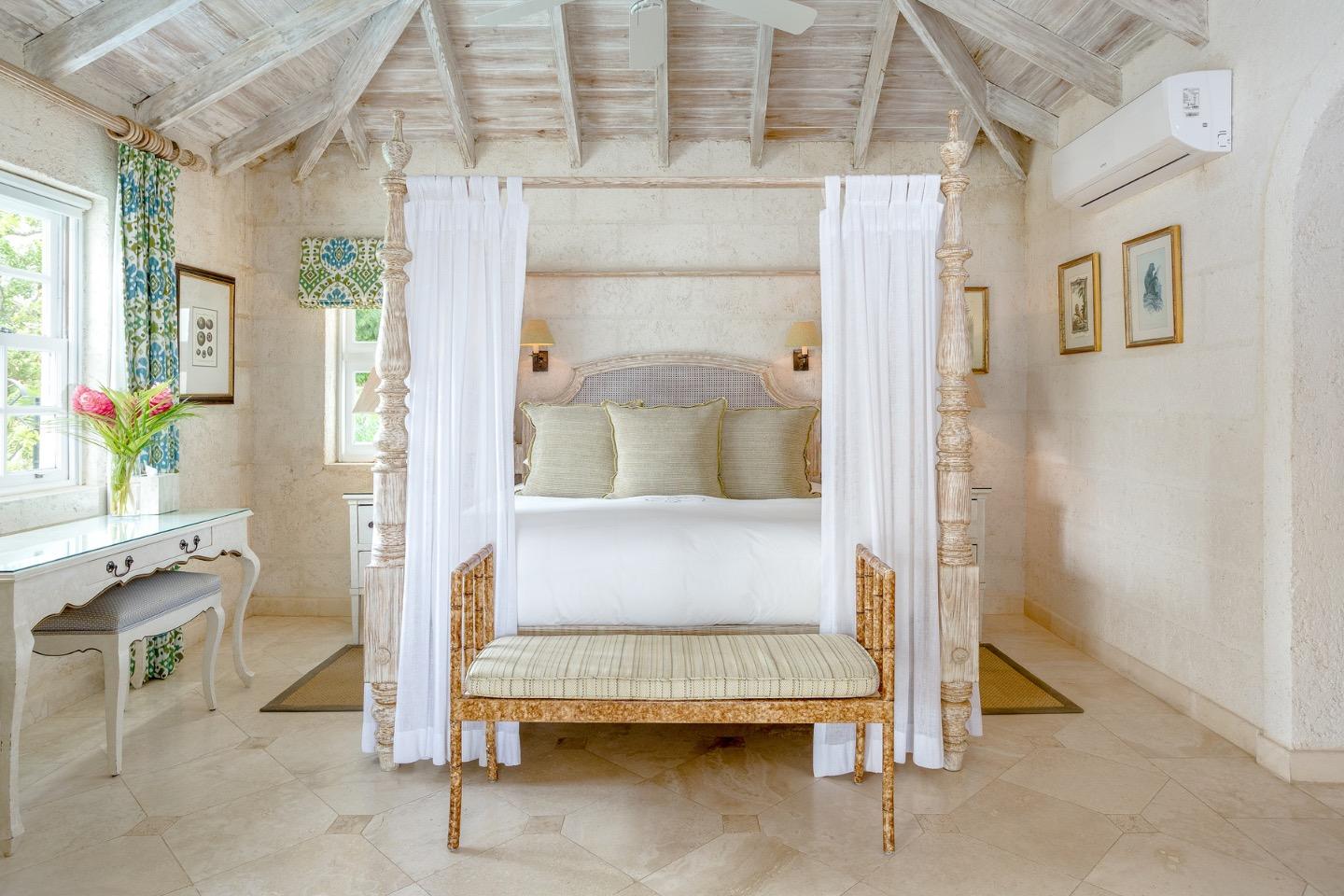 A four-poster bed hung with white curtains beneath a sloped wooden ceiling, with pastel pillows, a writing desk and fresh flowers to one side.