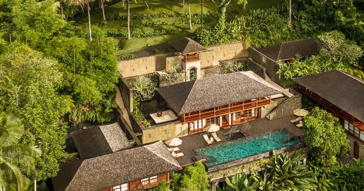 An aerial view of a thatched-roof hotel complex with a swimming pool, terraces and stone walls set among dense jungle.