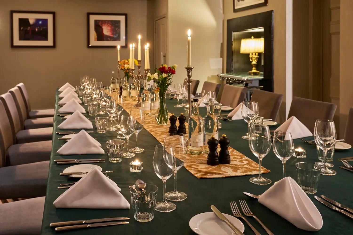 A long dining table with a green cloth, lit candles, folded white napkins, glasses and place settings laid for a private dinner.