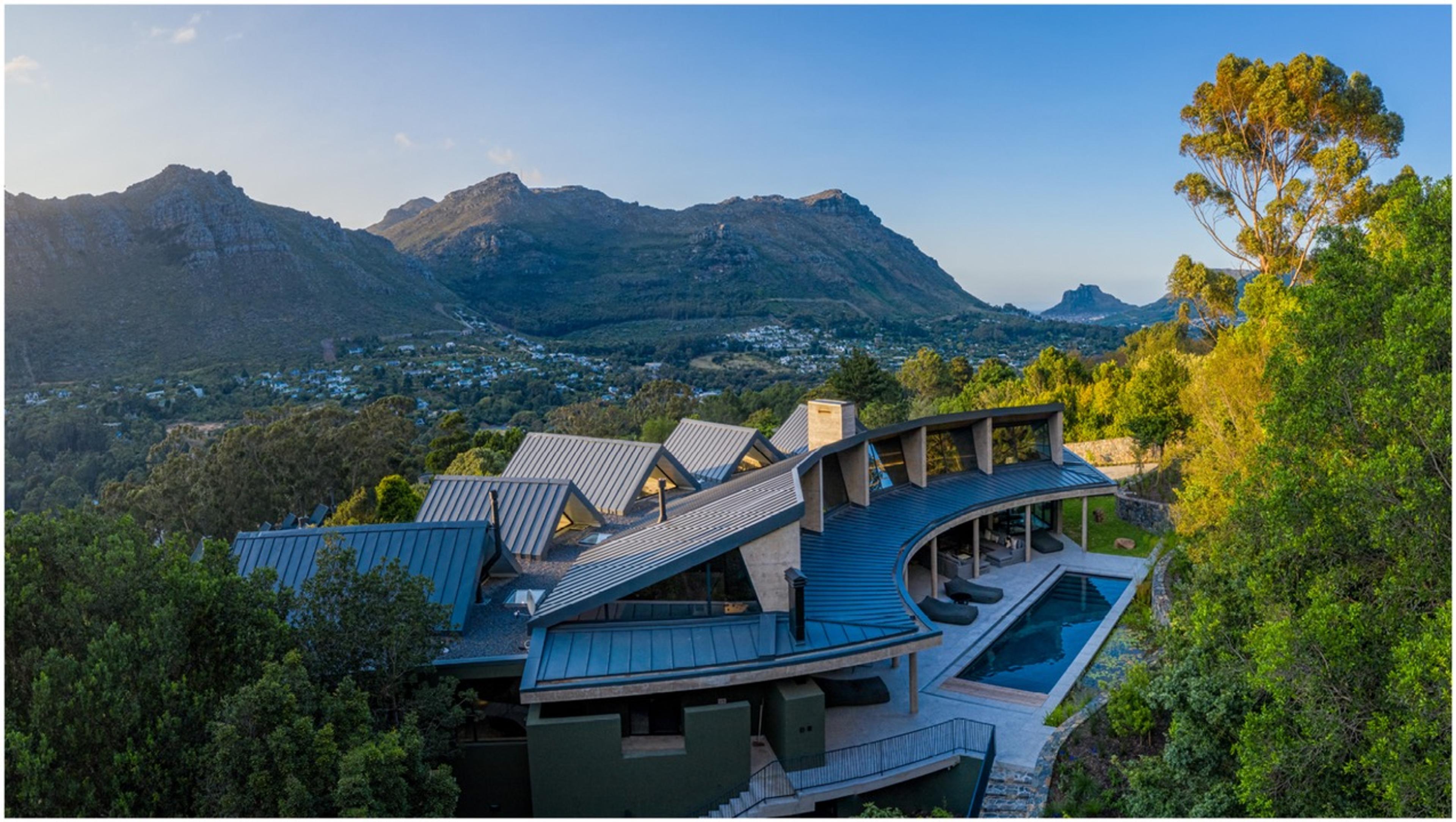 A long, curved building with dark gabled roofs and a swimming pool, built into a wooded hillside with mountains and a town in the valley beyond.