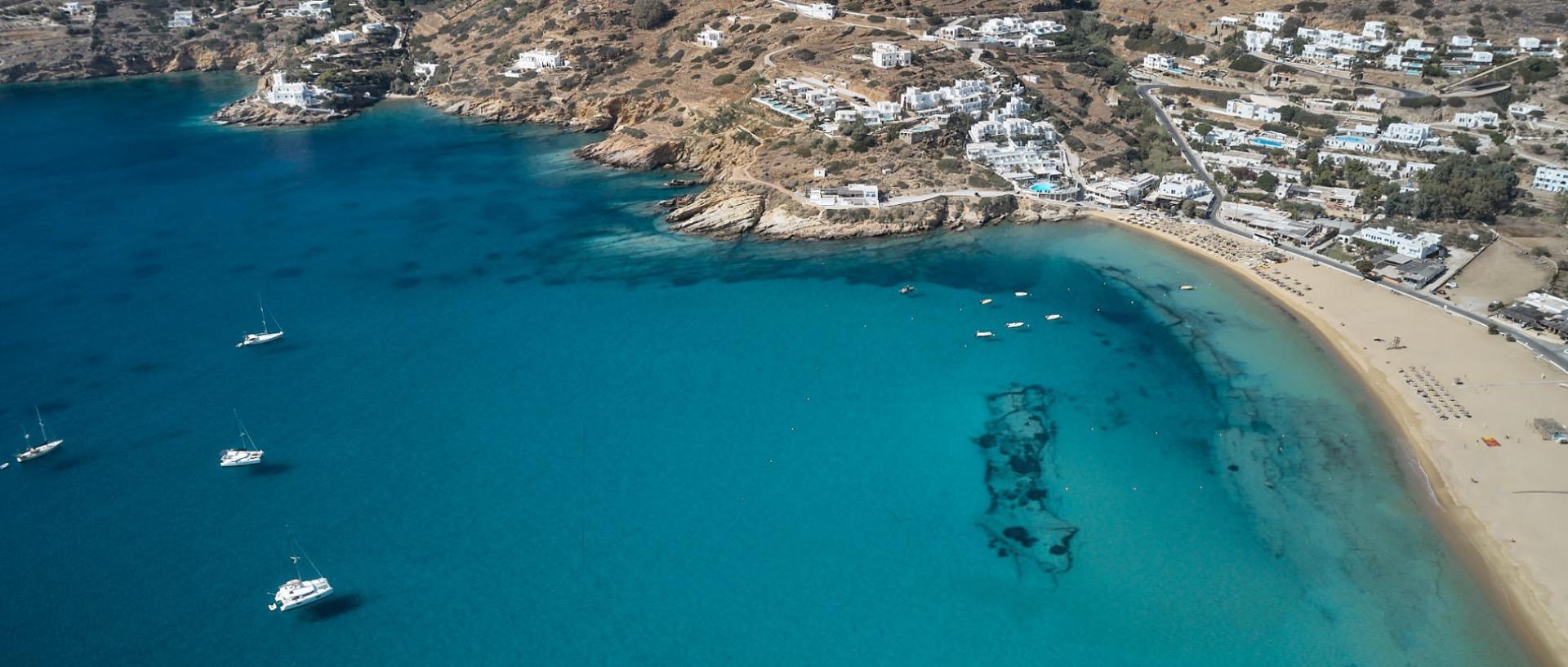 An aerial view of a bay with sailing boats, a sandy beach, a white village on the hillside, and the hotel's villas and pools on the headland.