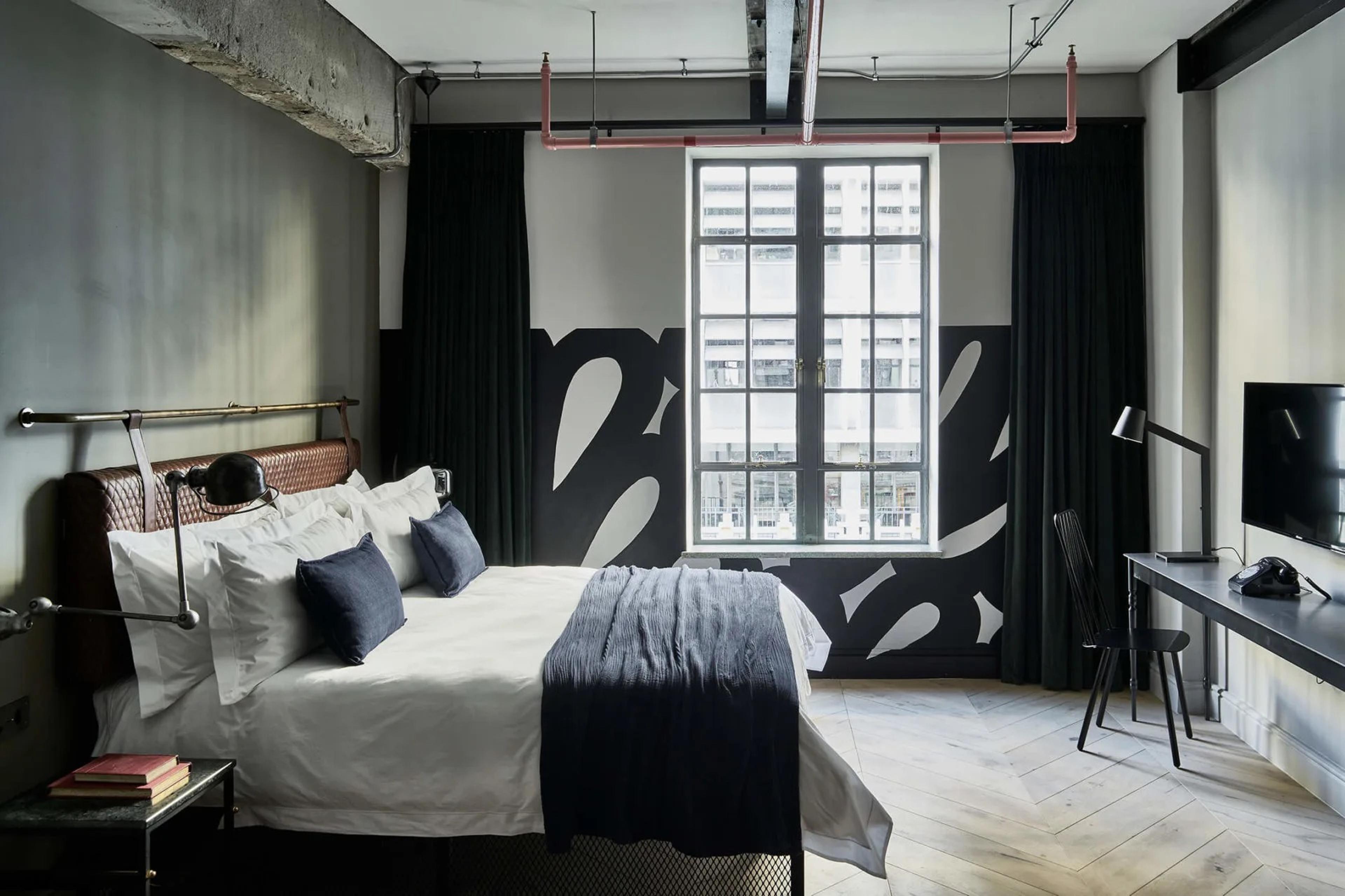 A double bed with white linen and dark blue cushions in a room with a black-and-white painted wall, a window onto the city, and a desk with a lamp and a wall-mounted television.