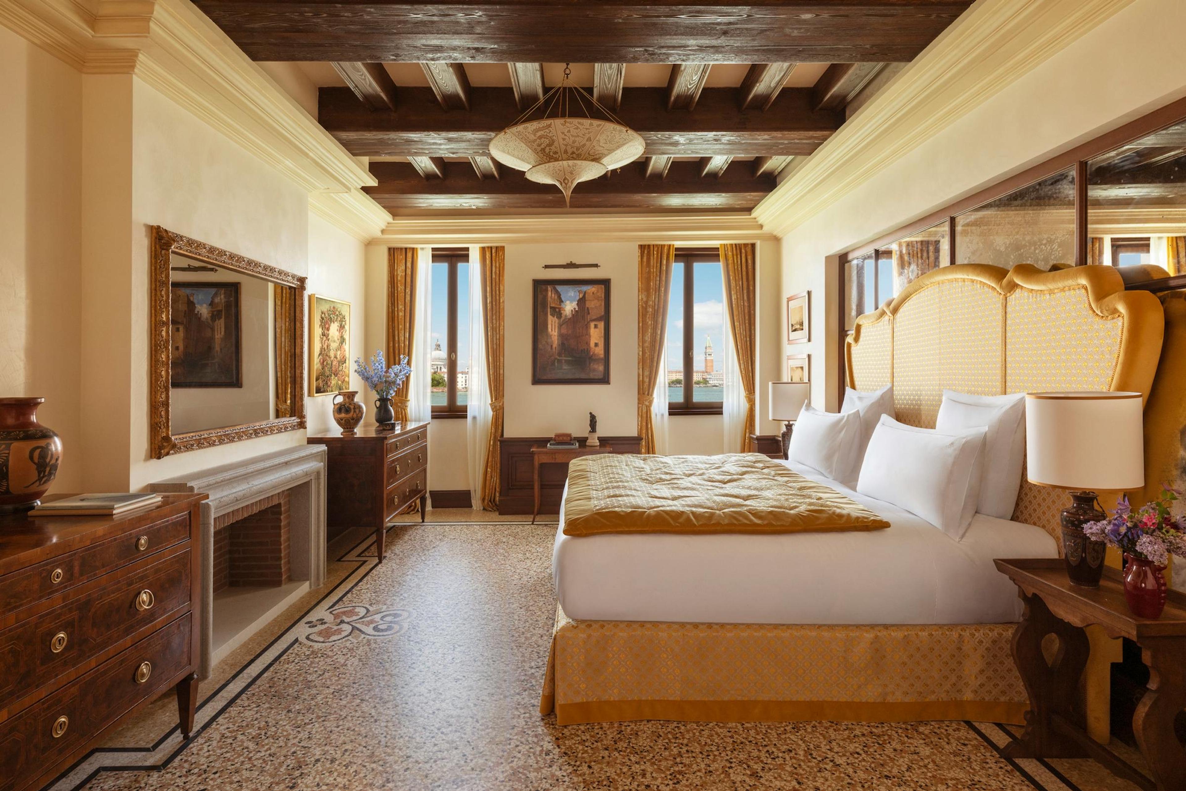 A bedroom with a gold upholstered headboard, a wood-beamed ceiling, a terrazzo floor, and two curtained windows looking out over Venice rooftops and a campanile.