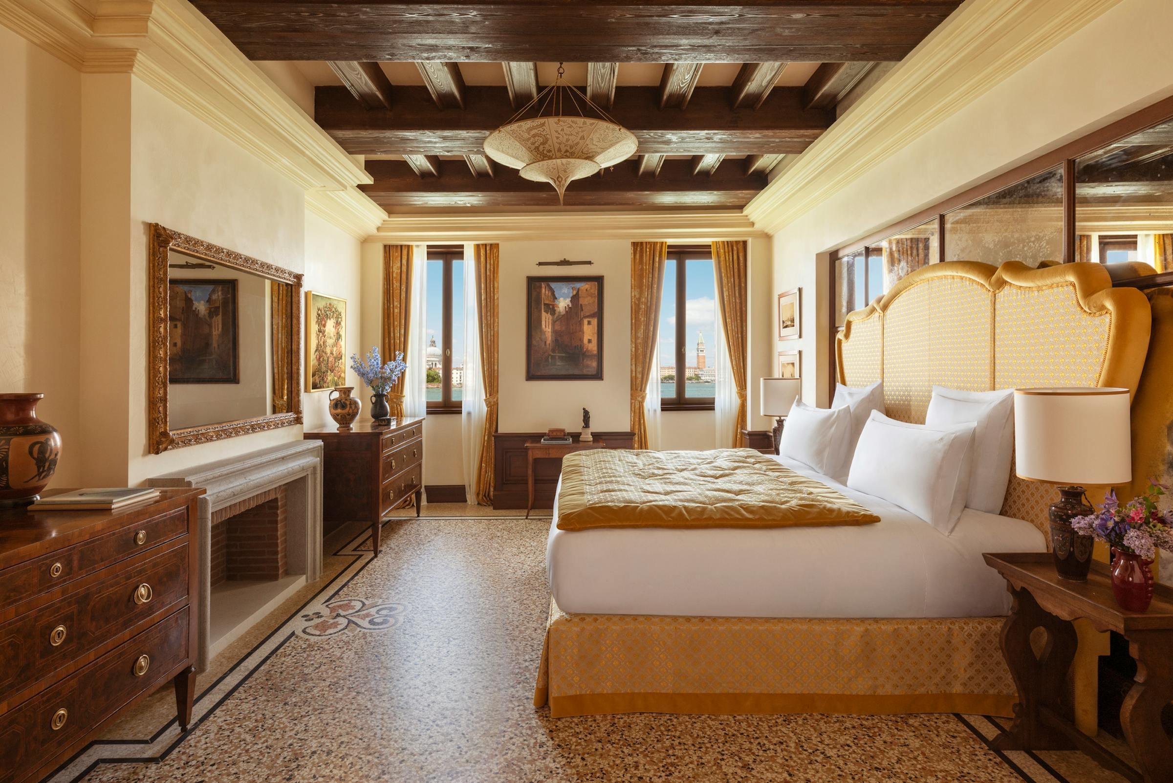 A bedroom with a gold upholstered headboard, a wood-beamed ceiling, a terrazzo floor, and two curtained windows looking out over Venice rooftops and a campanile.