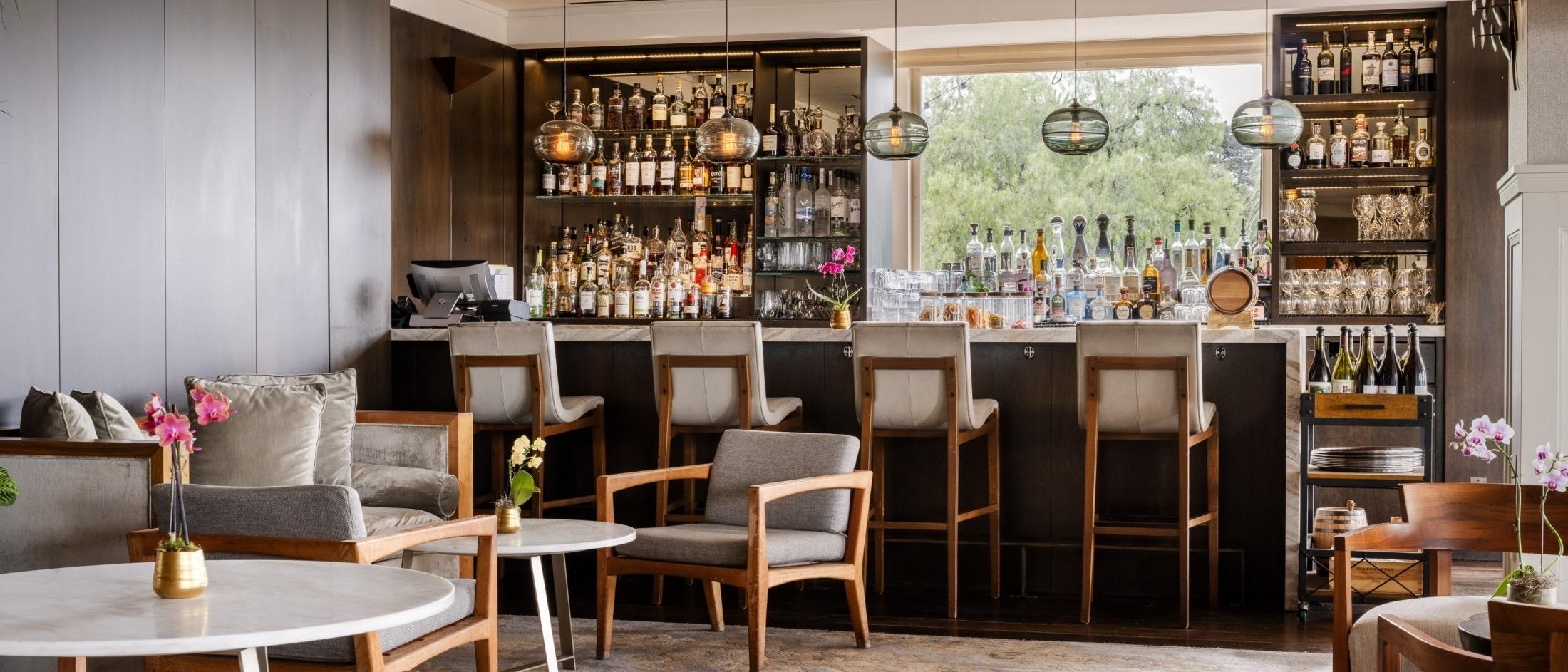A hotel bar with wooden stools along a counter backed by shelves of bottles, pendant lights, and a seating area with armchairs and a marble table in the foreground.