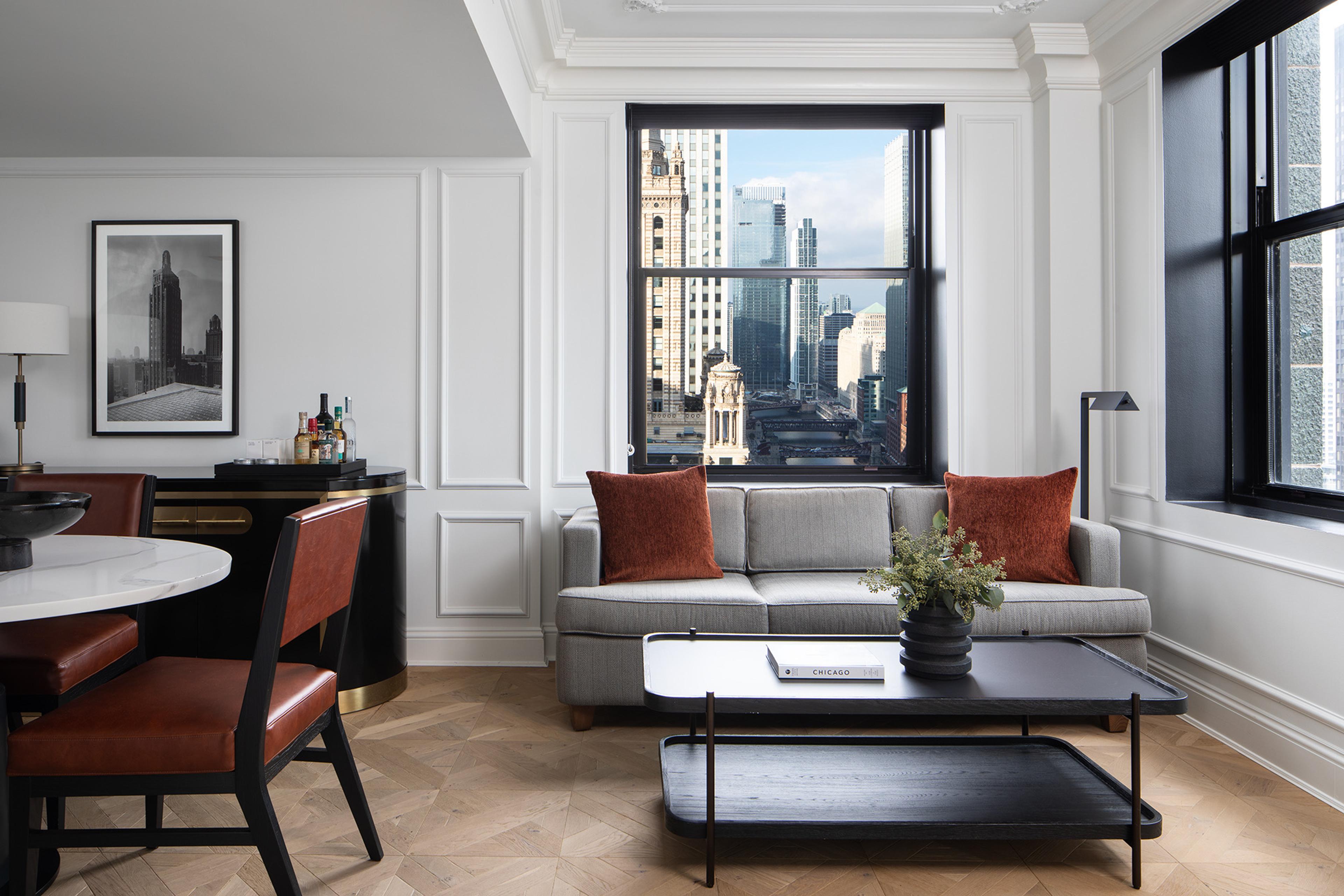 A hotel suite living and dining area with a round marble table, leather chairs, a red credenza, and a grey sofa, with a view of nearby towers through the window.