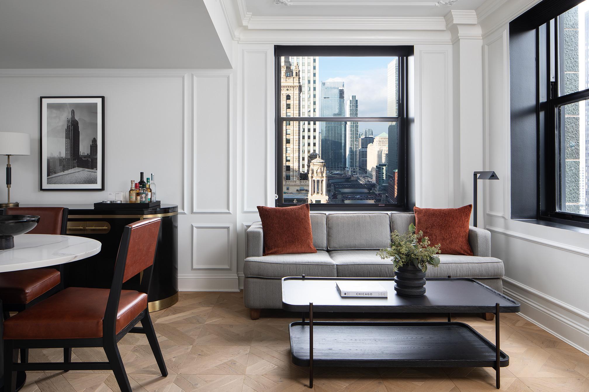 A hotel suite living and dining area with a round marble table, leather chairs, a red credenza, and a grey sofa, with a view of nearby towers through the window.