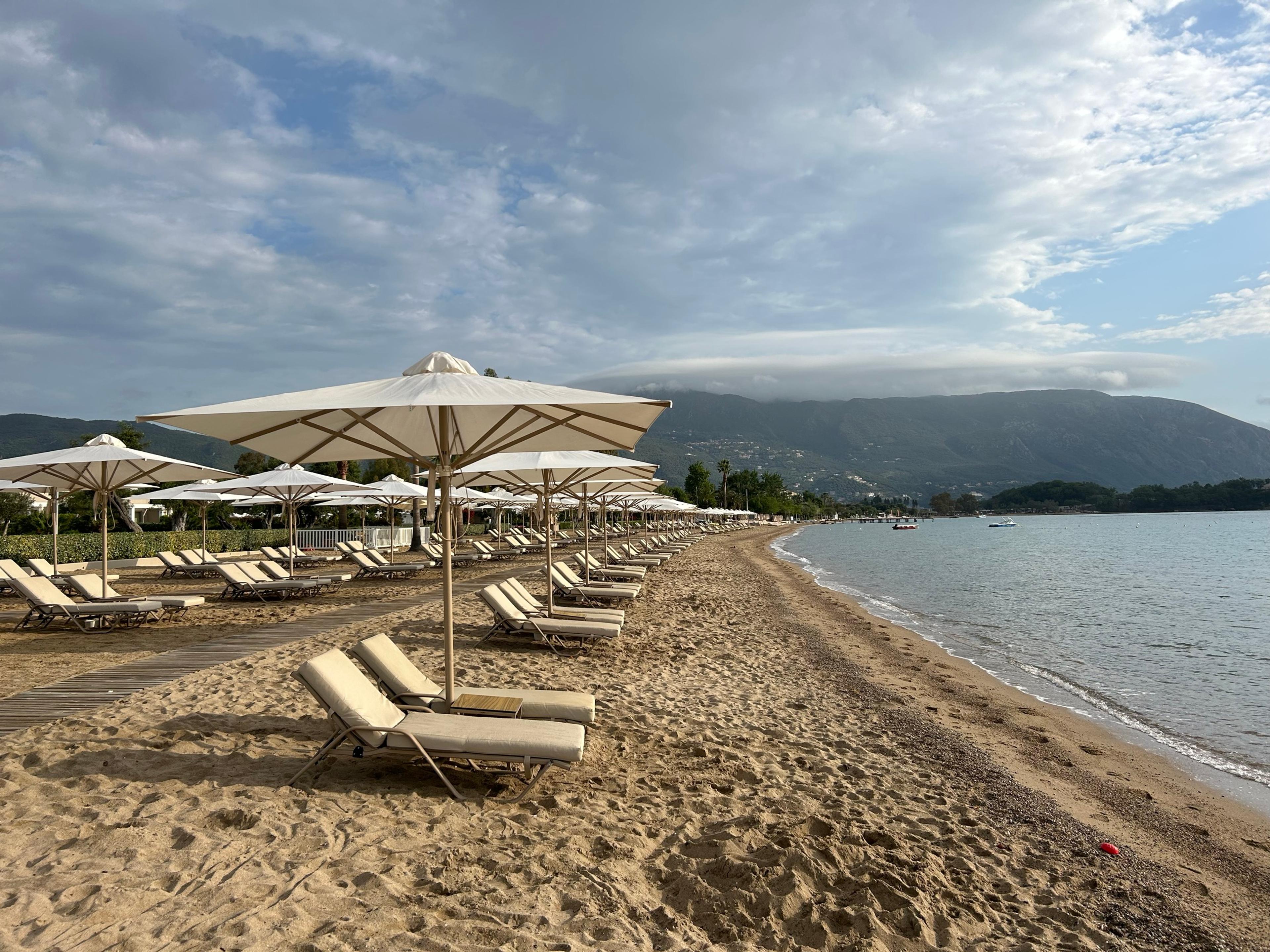 A sandy beach at Dassia lined with parasols and sun loungers, calm water and wooded hills under a cloudy sky