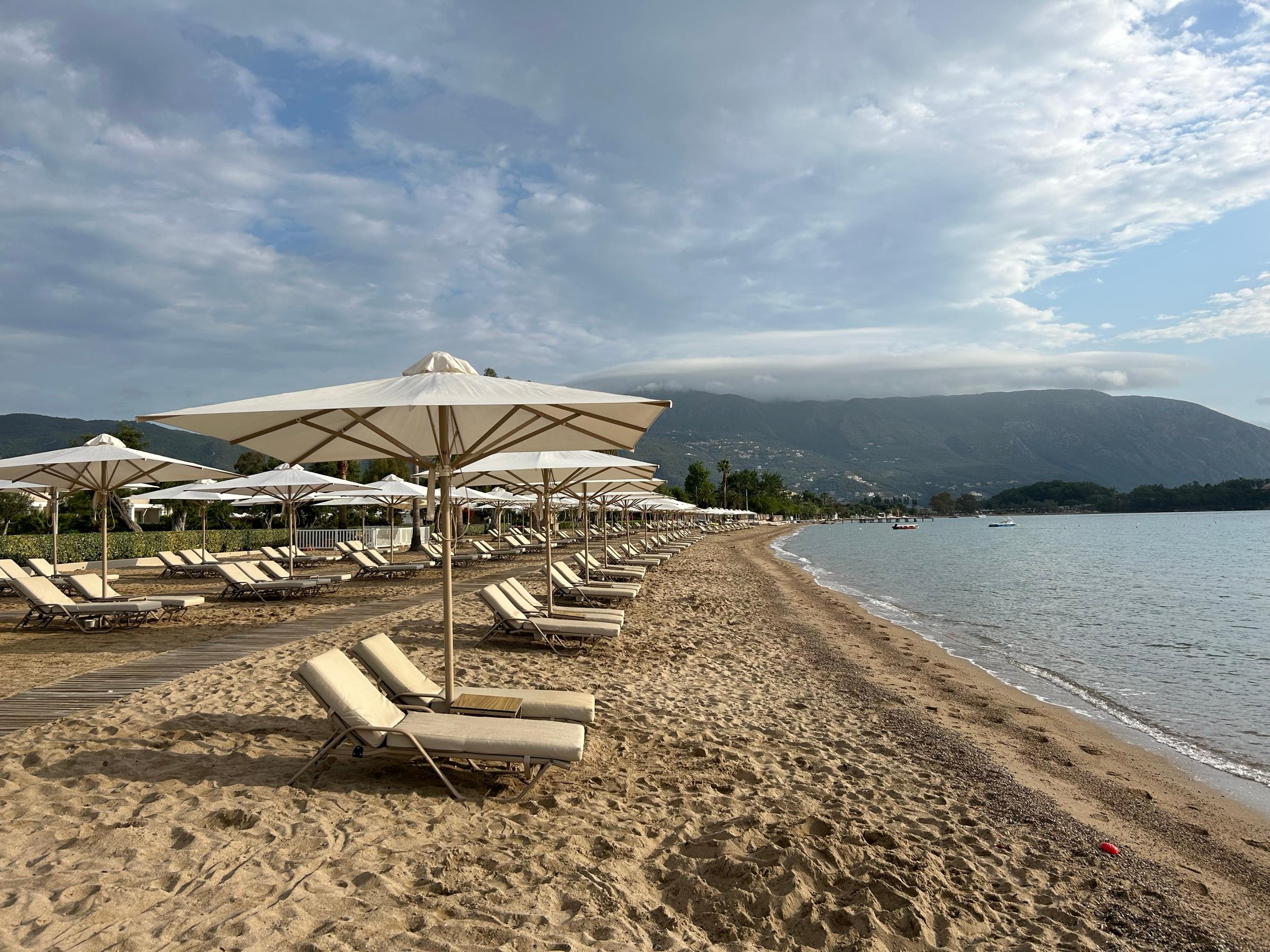 A sandy beach at Dassia lined with parasols and sun loungers, calm water and wooded hills under a cloudy sky