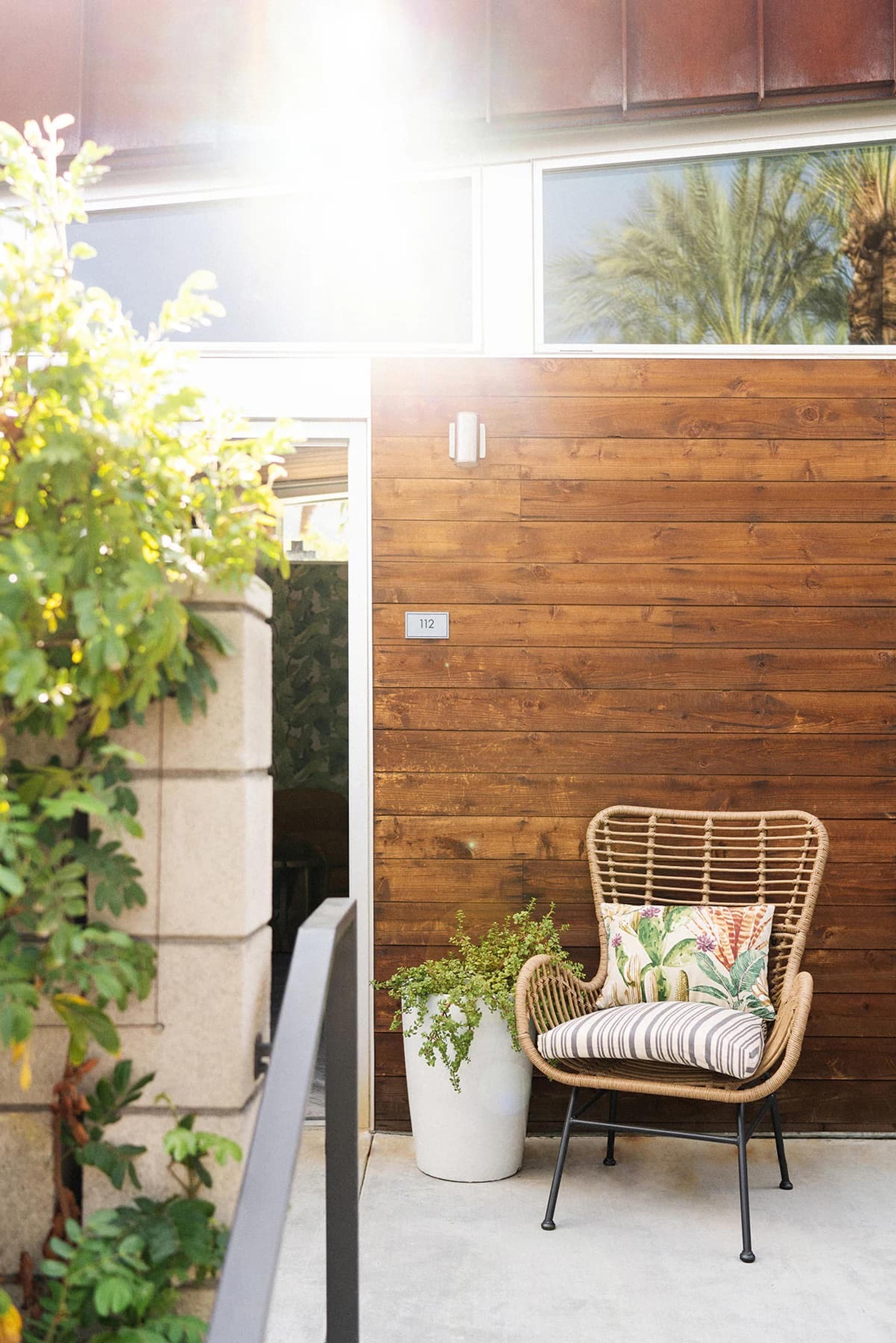 A timber-clad wall beside an open glass doorway, with a wicker armchair holding a striped and patterned cushion, a white planter, and a small plaque with the number 112.