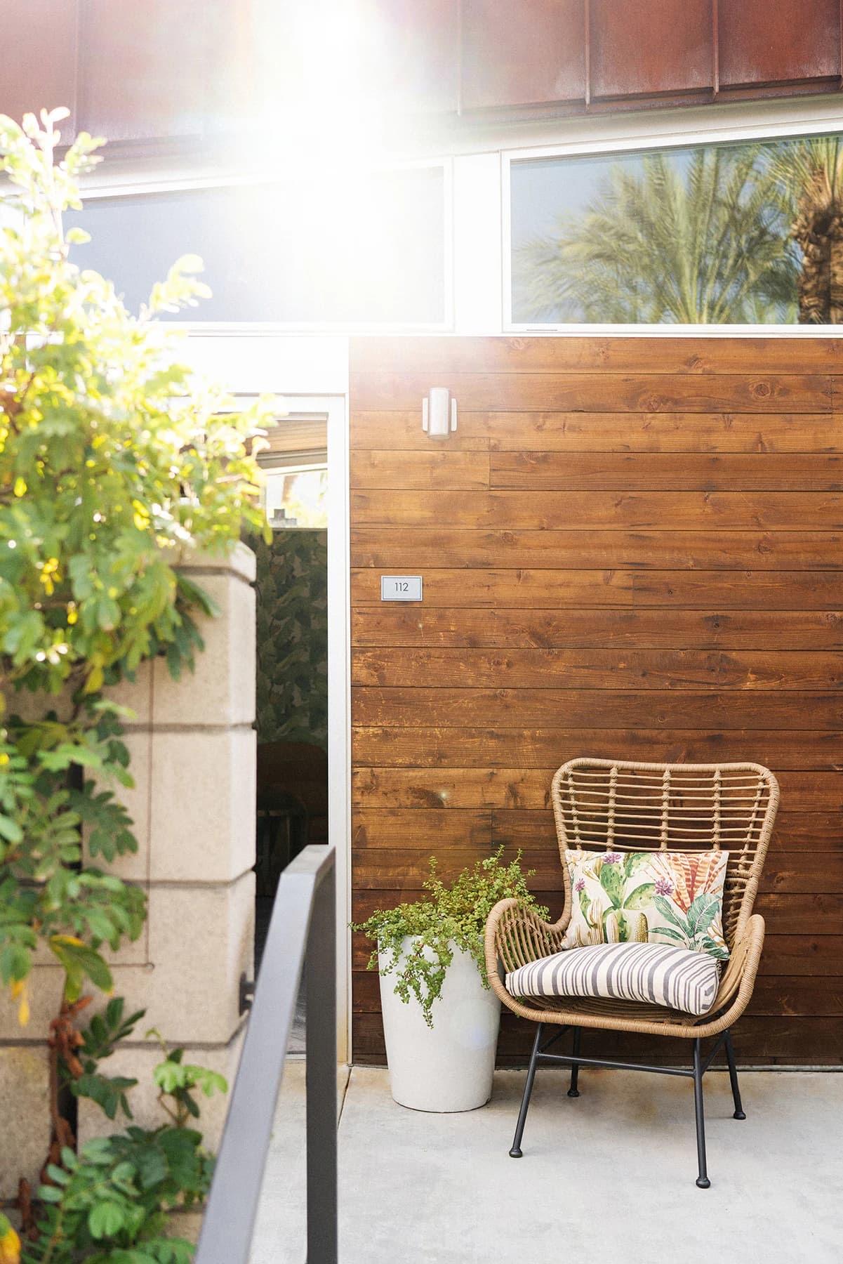 A timber-clad wall beside an open glass doorway, with a wicker armchair holding a striped and patterned cushion, a white planter, and a small plaque with the number 112.