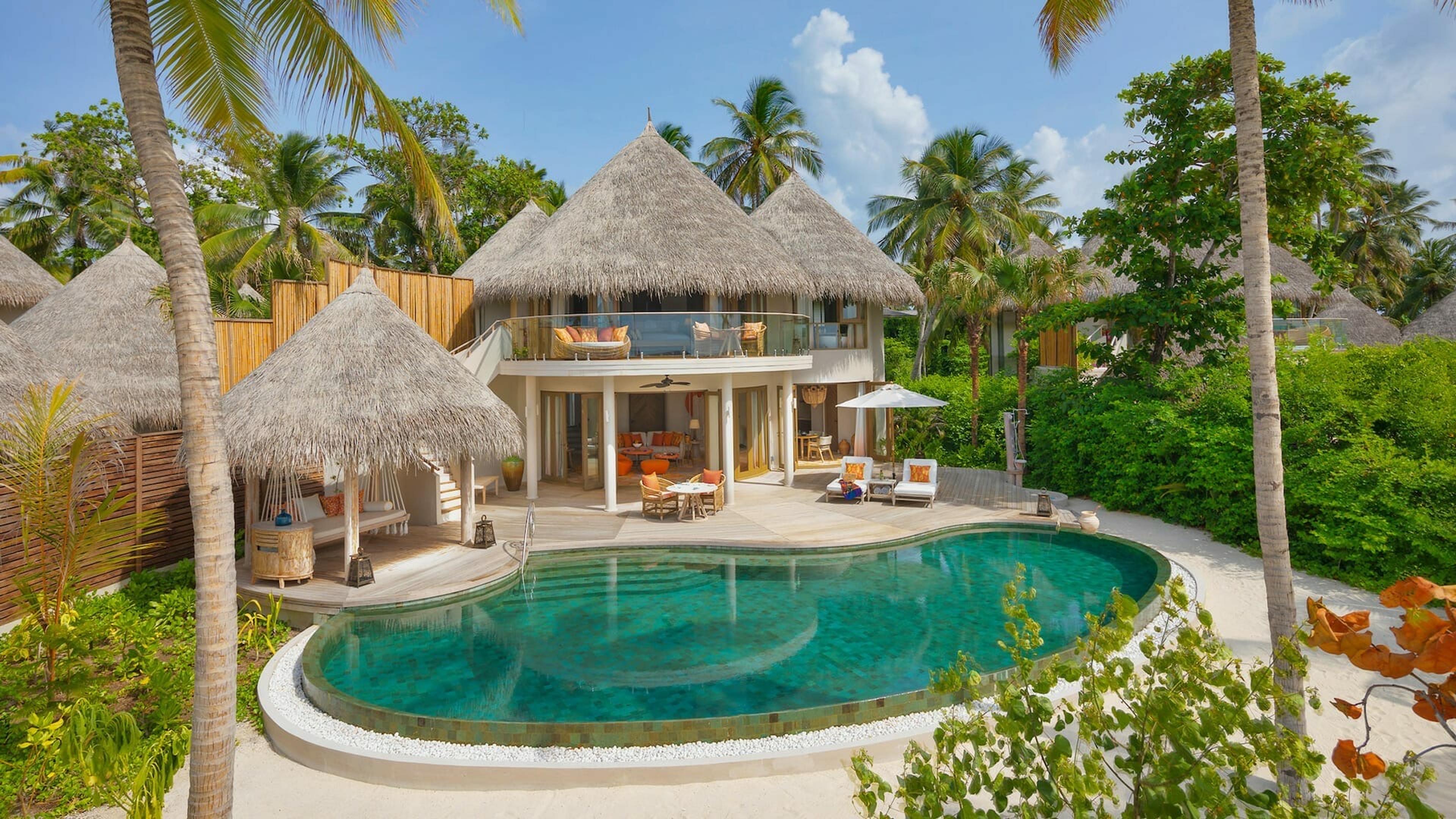 A two-storey thatched villa with a large curved pool, outdoor dining area and a day bed pavilion, surrounded by palm trees.
