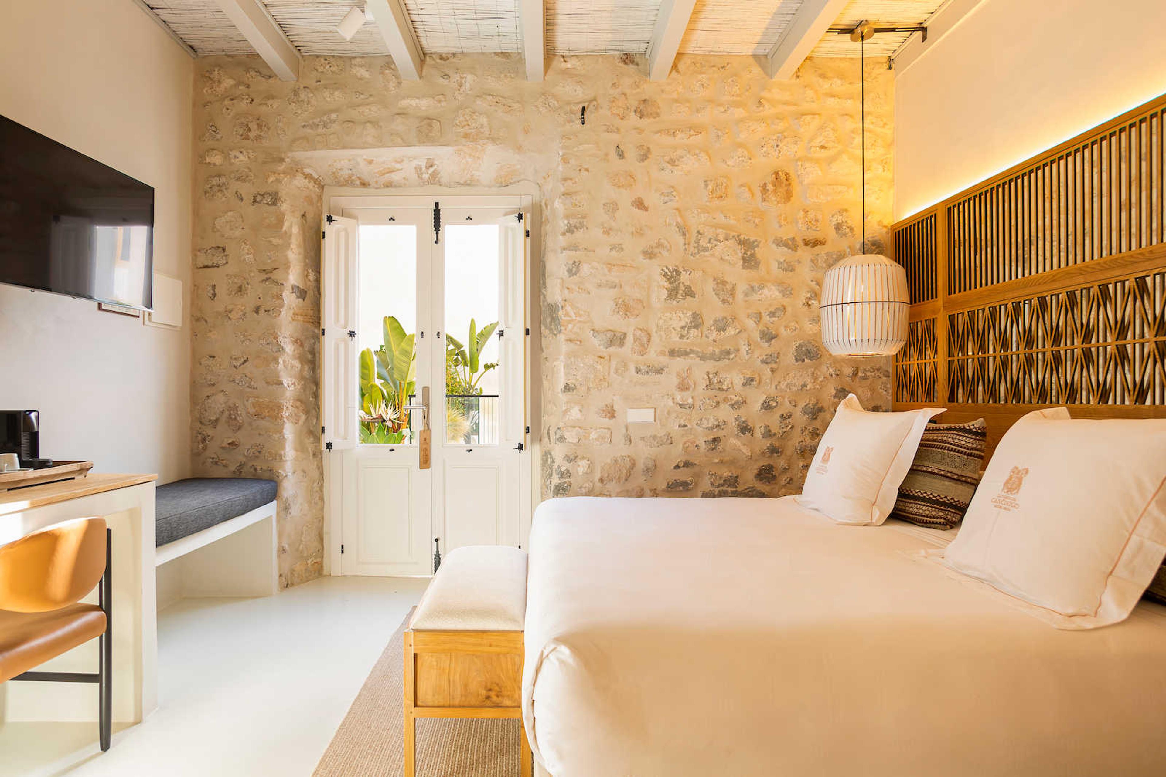 A bedroom with a stone wall and a bamboo headboard, its white doors open onto a terrace with banana plants, and a desk and wall television on the left.