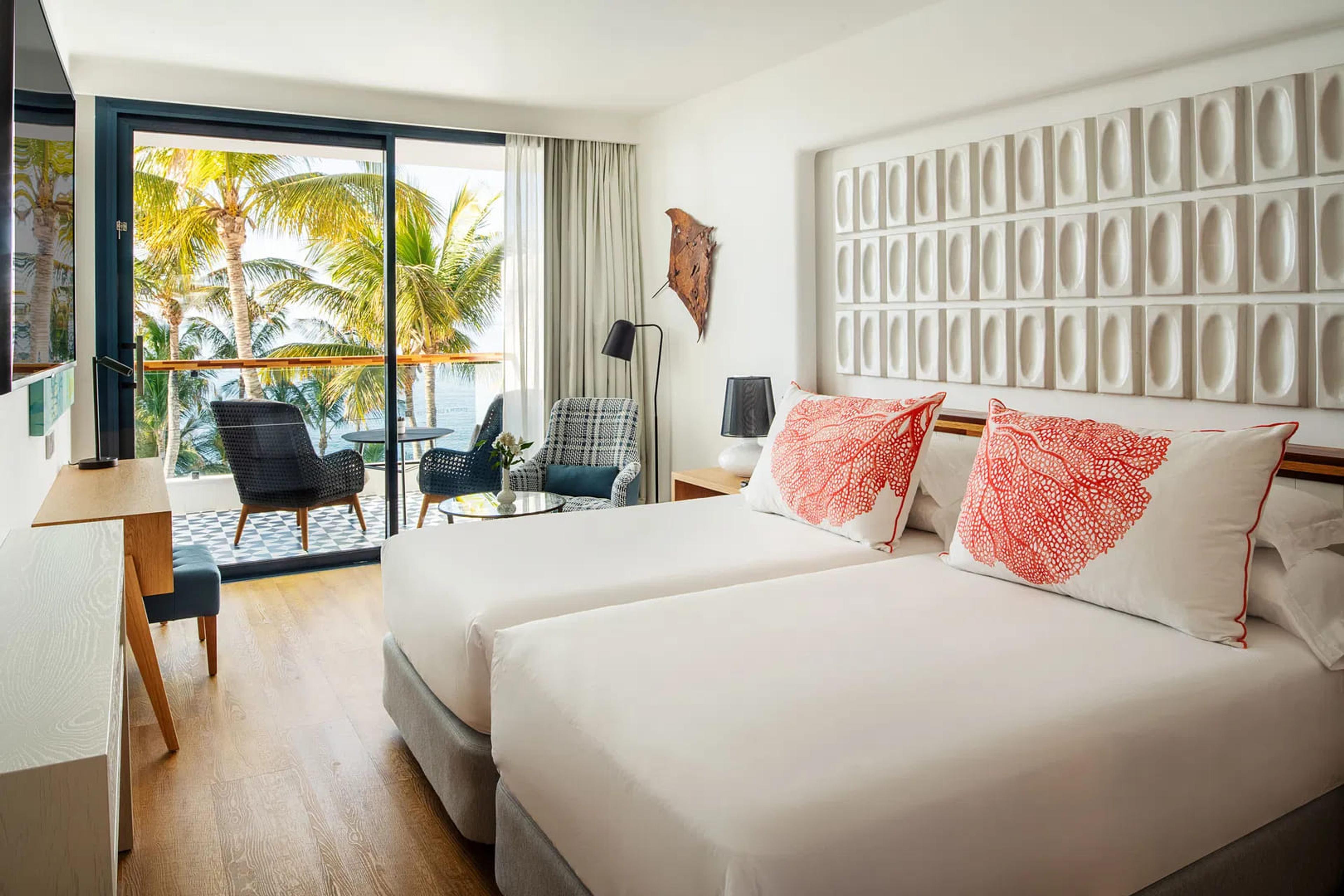 A twin room with coral patterned cushions against a textured white headboard, a desk and seating area leading to a balcony with palm trees beyond.