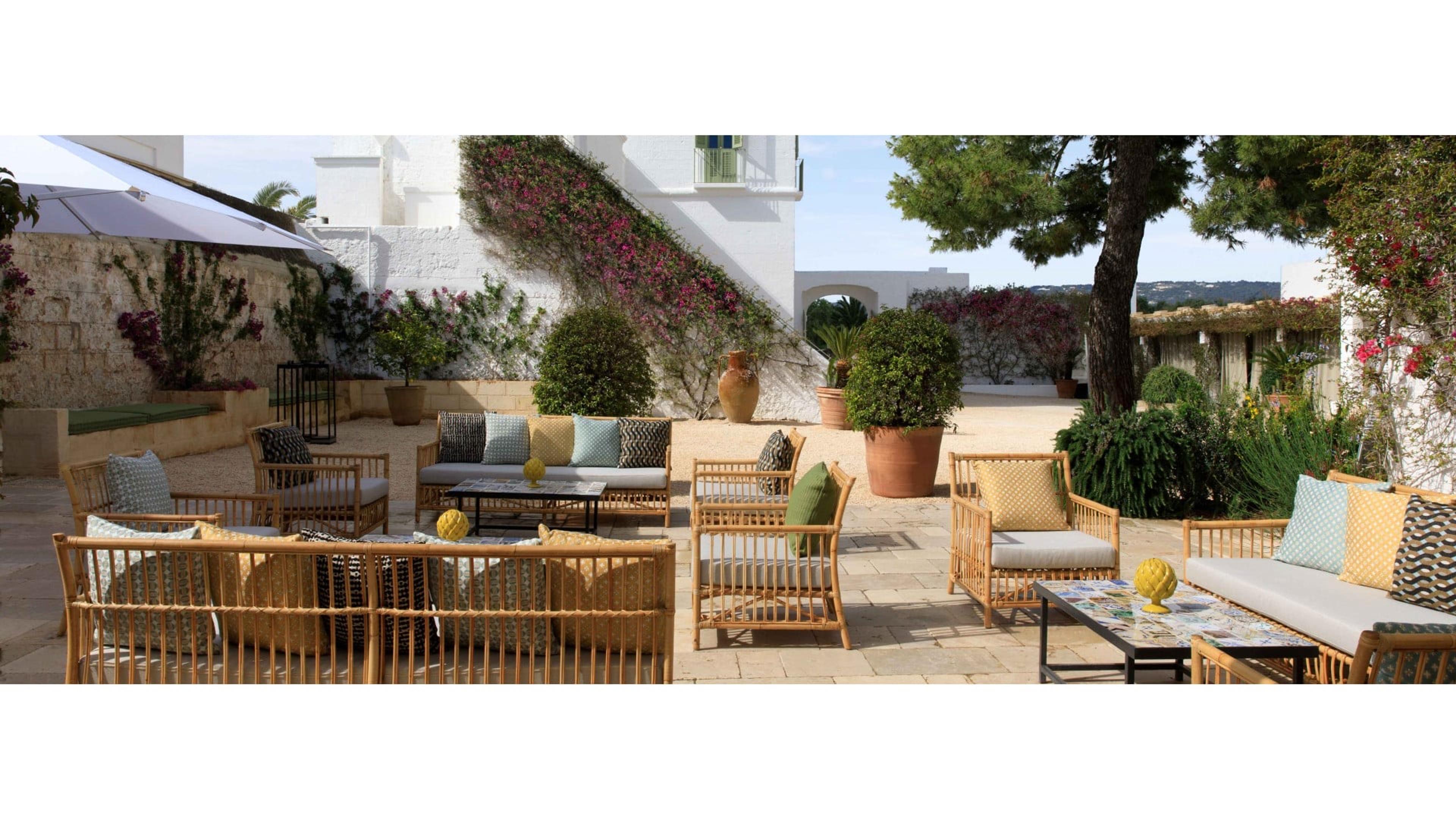 A paved courtyard with rattan sofas and armchairs around low tables, bougainvillea climbing a whitewashed wall beside a pine tree and open countryside beyond.