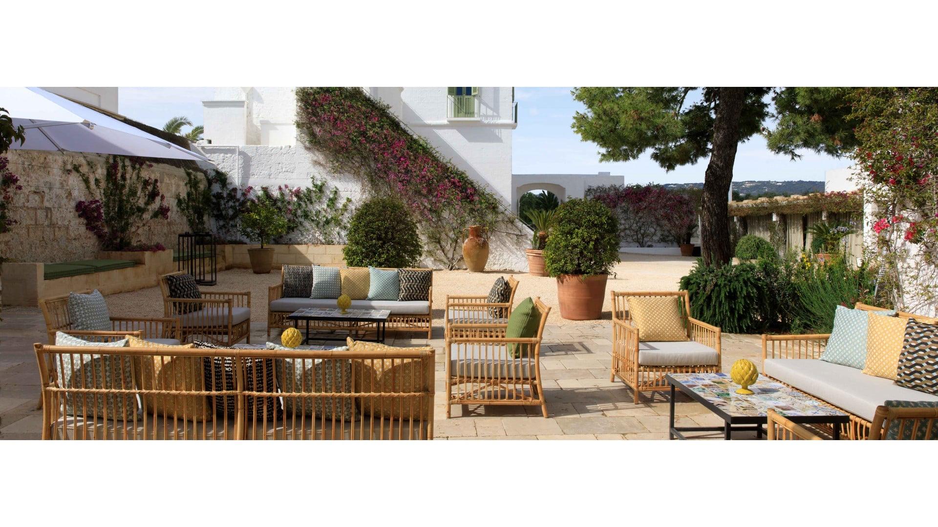A paved courtyard with rattan sofas and armchairs around low tables, bougainvillea climbing a whitewashed wall beside a pine tree and open countryside beyond.