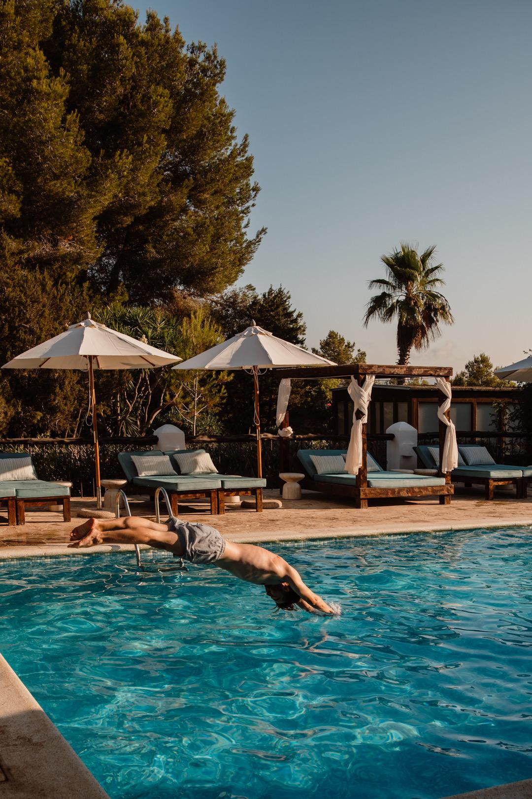 A man dives into a swimming pool past a row of white parasols and cushioned loungers.