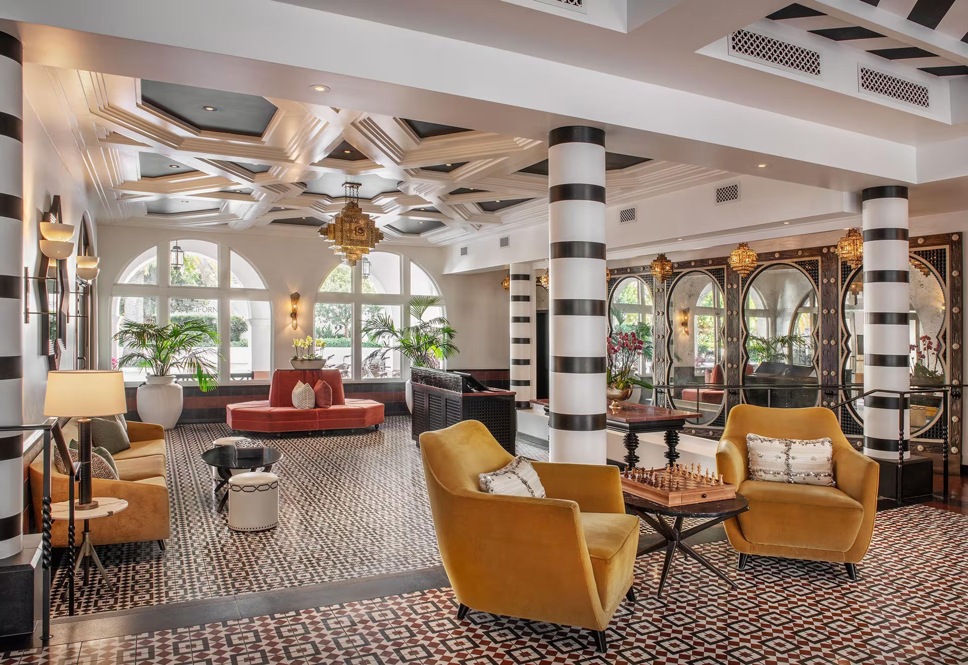 A hotel lobby with black and white striped columns, a patterned tile floor, mustard velvet armchairs around a chess table, and arched mirrored panels along one wall.