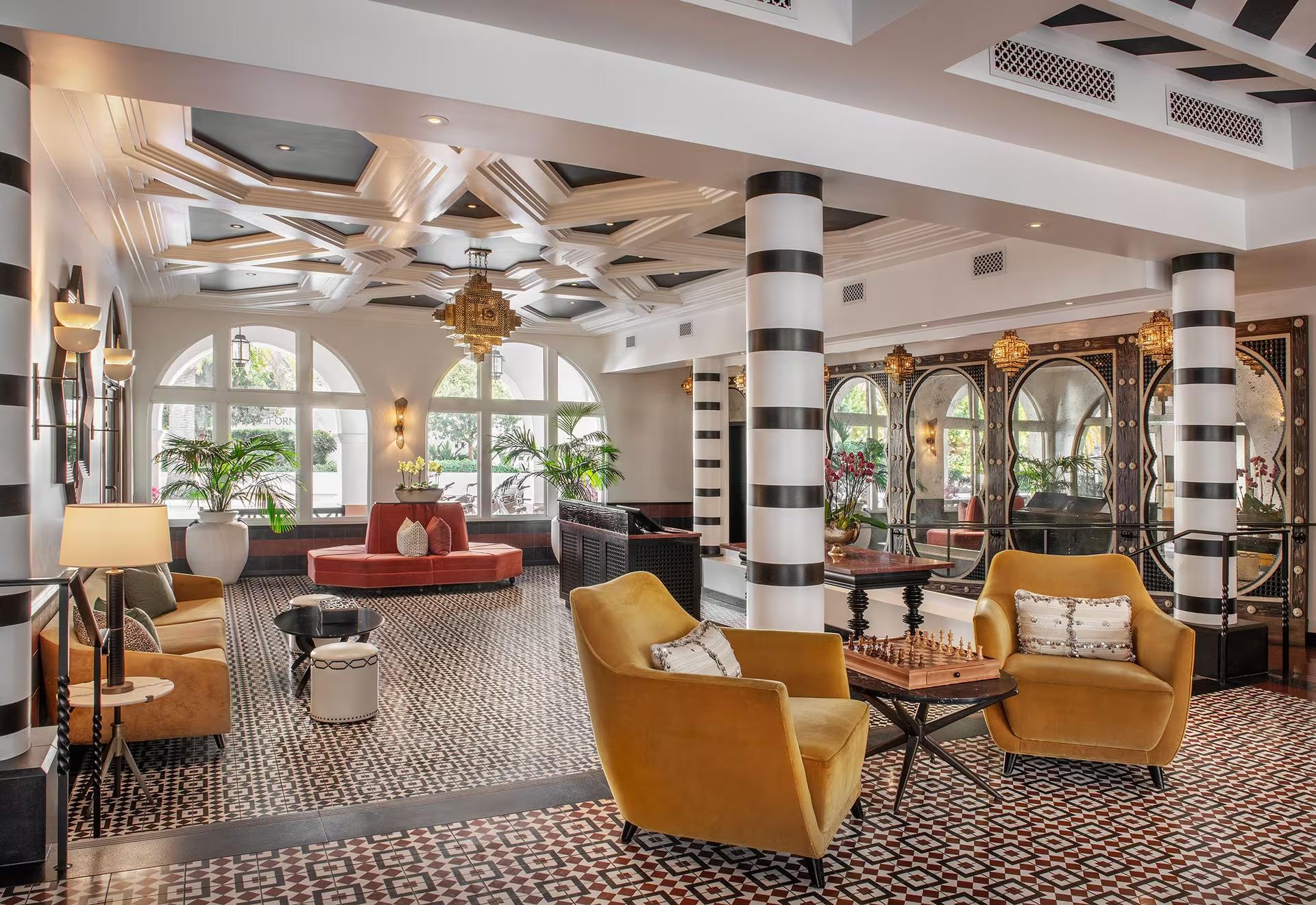 A hotel lobby with black and white striped columns, a patterned tile floor, mustard velvet armchairs around a chess table, and arched mirrored panels along one wall.