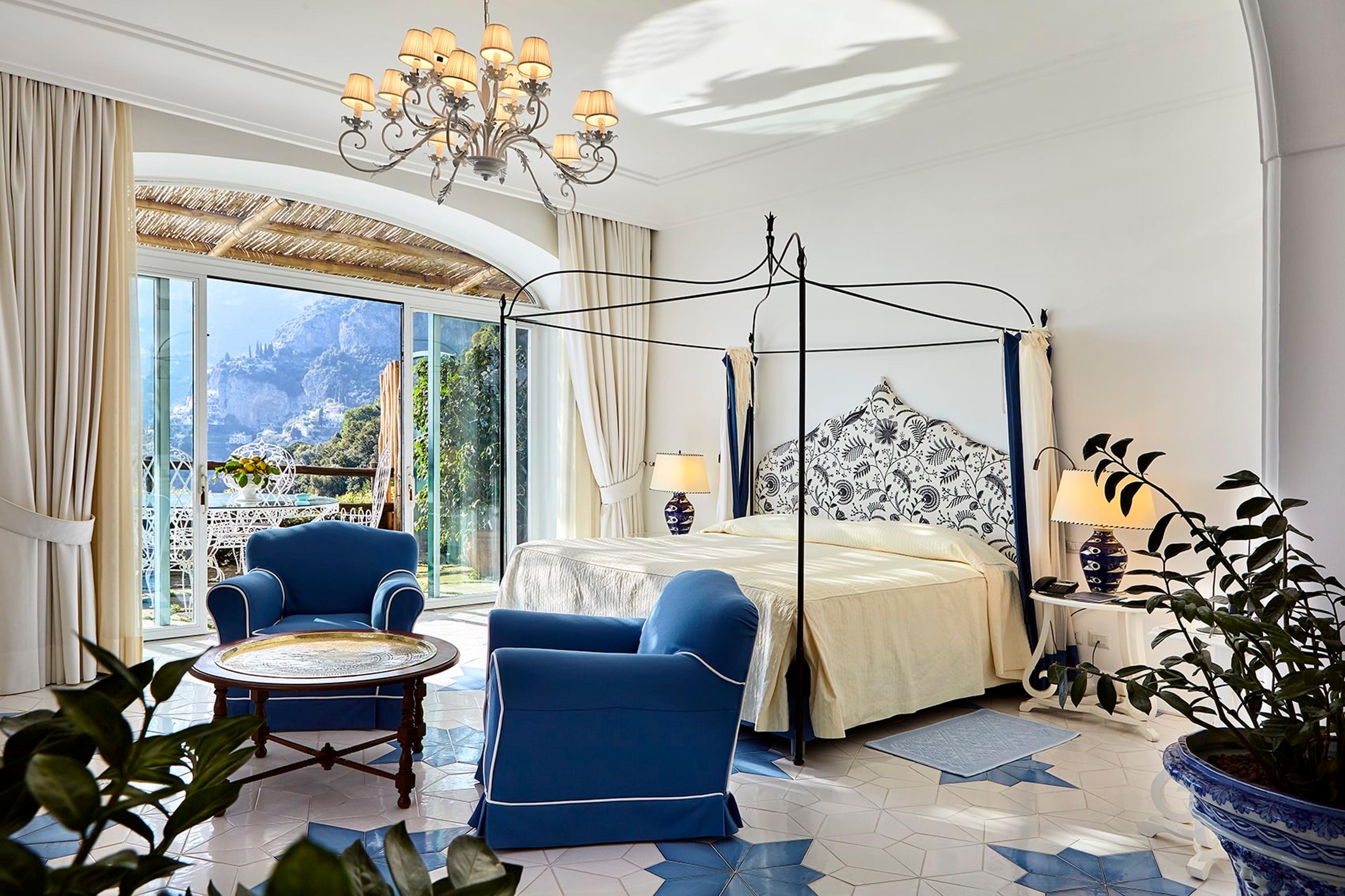 A hotel bedroom with a wrought-iron four-poster bed, two blue armchairs, a patterned tile floor, and an arched window looking over the coast.