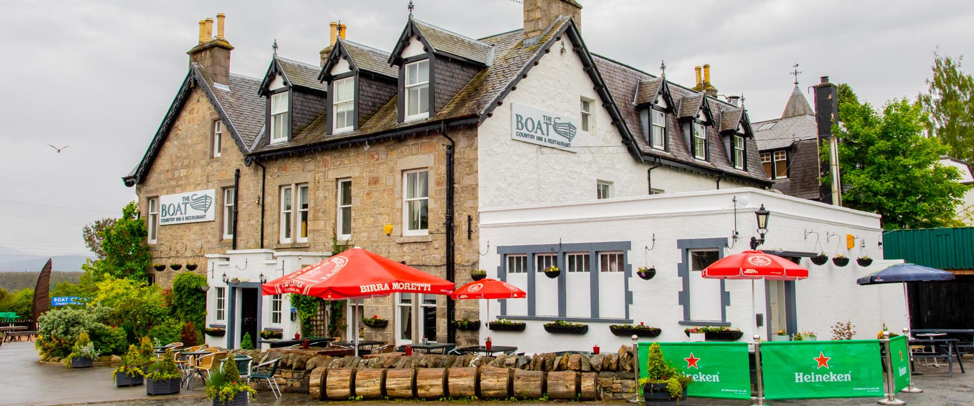 A stone and white painted inn with gabled dormers, red parasols advertising Birra Moretti shading outdoor tables, and Heineken branded barriers along the pavement.