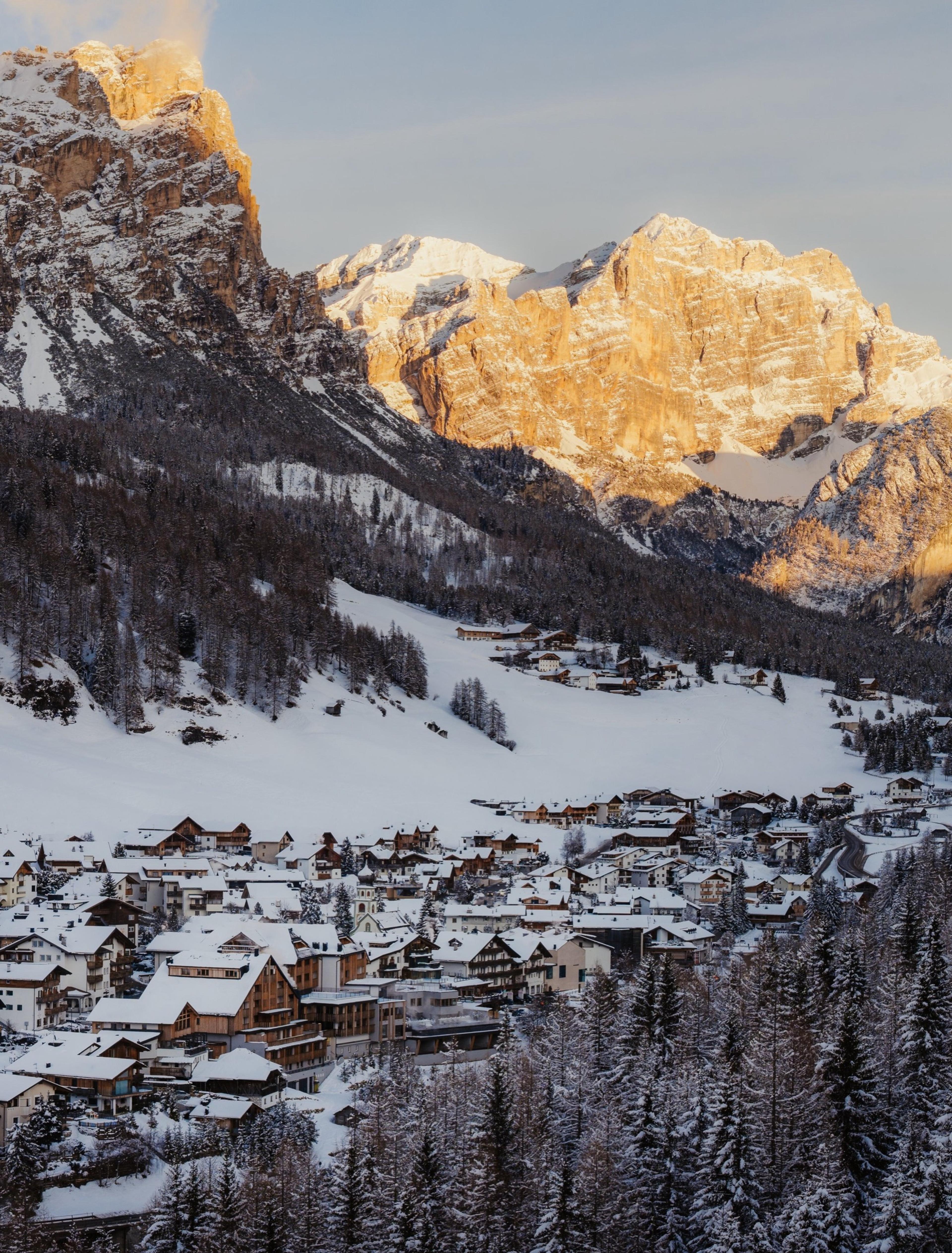 a mountain village under snow covered Dolomite peaks lit gold at dusk, with dark pine forest and rooftops below