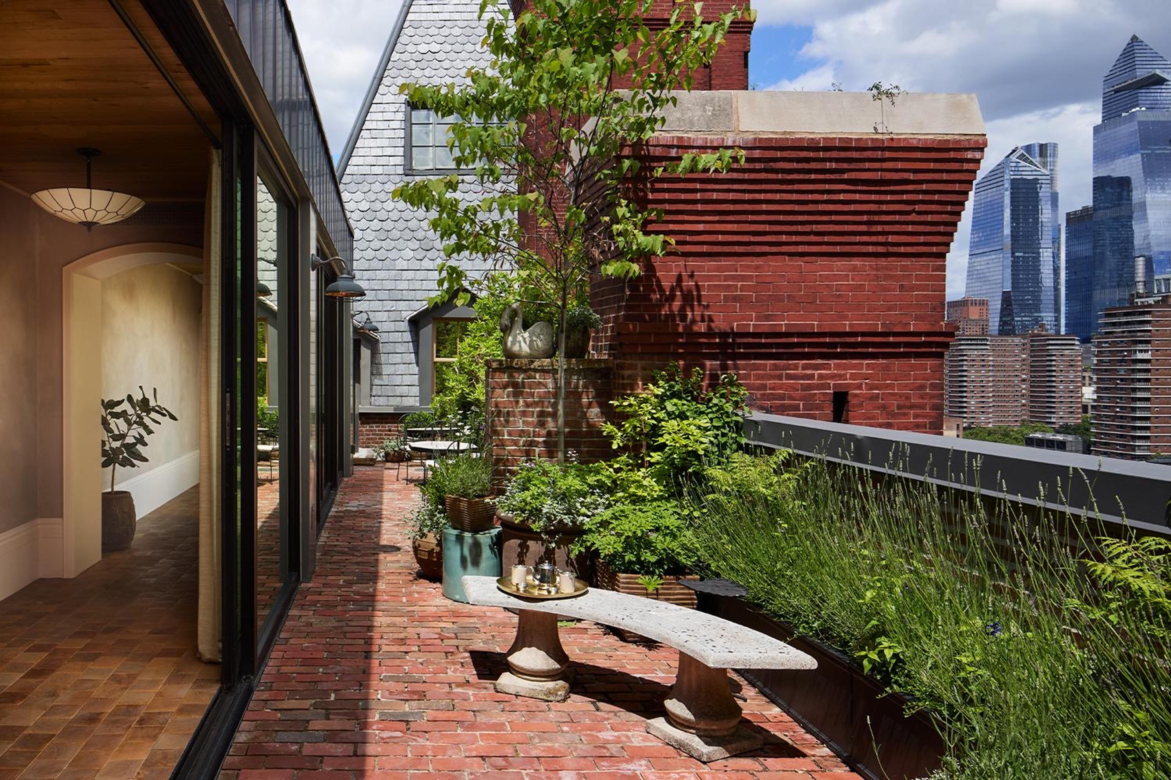 A rooftop terrace with potted trees and shrubs, a small table set for coffee, and the Manhattan skyline including the Empire State Building beyond.