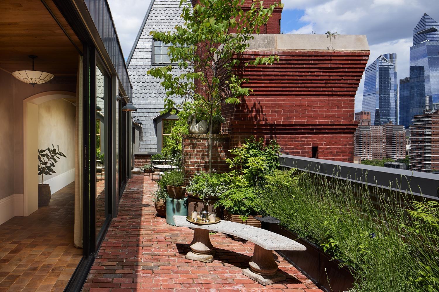 A rooftop terrace with potted trees and shrubs, a small table set for coffee, and the Manhattan skyline including the Empire State Building beyond.