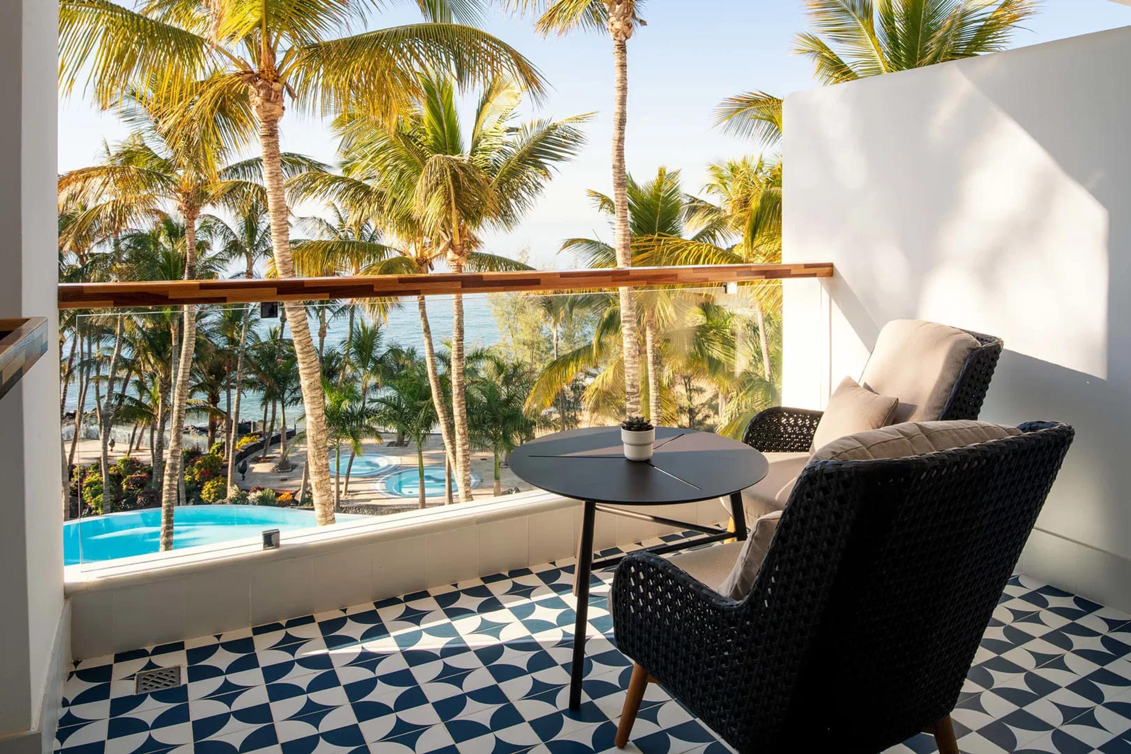 A tiled balcony with two woven armchairs and a small table looks out over swimming pools and rows of palm trees to the sea.