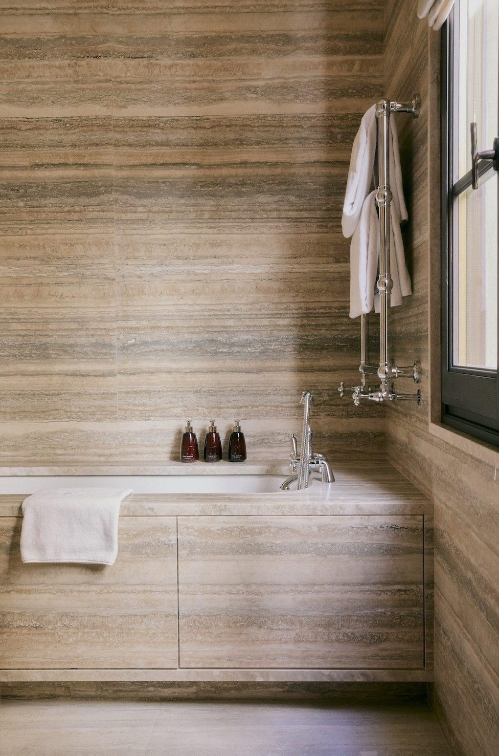 A travertine-clad bathroom with a built-in tub, a wall-mounted tap, folded towels on a rail, and a window at the side.