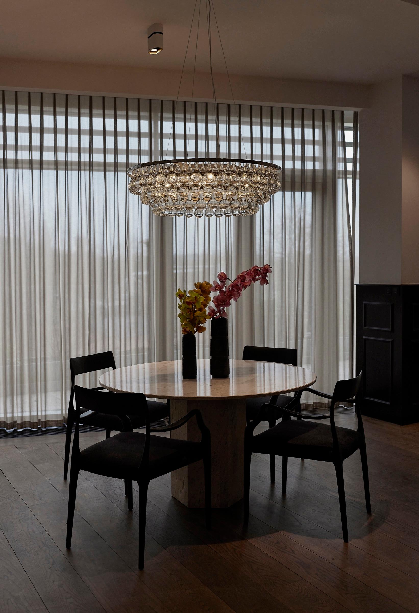A round travertine table set with orchids and four black chairs sits beneath a crystal chandelier, in front of floor-to-ceiling sheer curtains with a view of rooftops beyond.