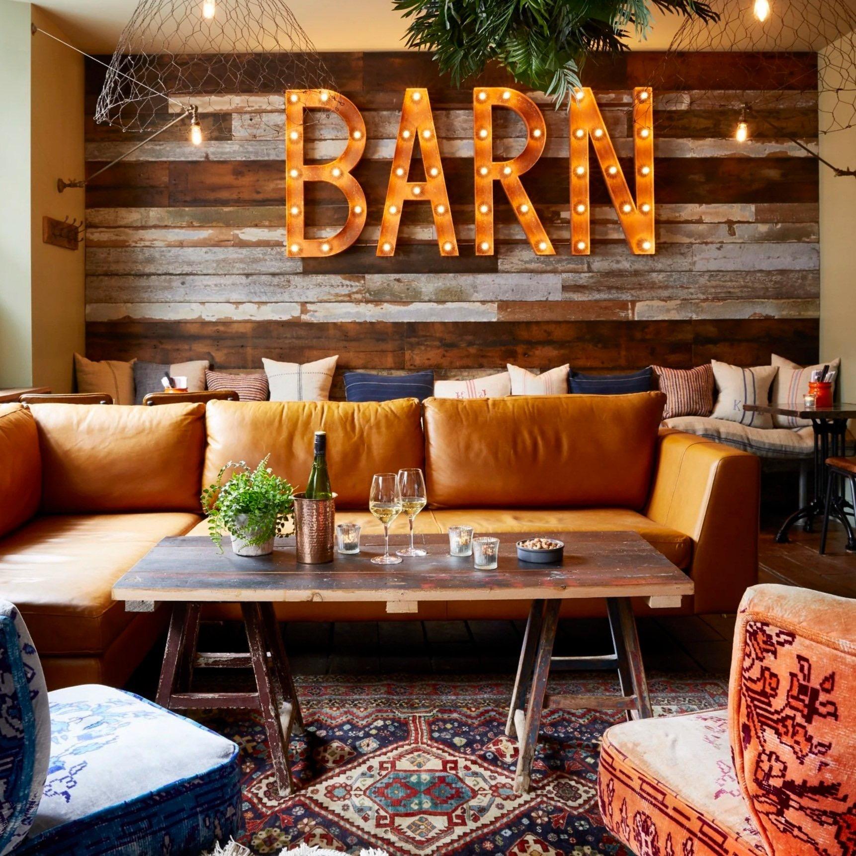 A lounge with a tan leather sofa and patterned armchairs around a low wooden table set with wine glasses, below a wall sign spelling BARN in light bulbs.