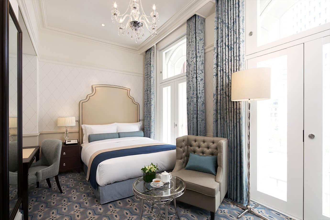A hotel bedroom with a crystal chandelier, a bed with a navy and white runner, a tufted beige headboard, a grey wingback armchair, a round side table with a tea set and flowers, and tall curtained windows.
