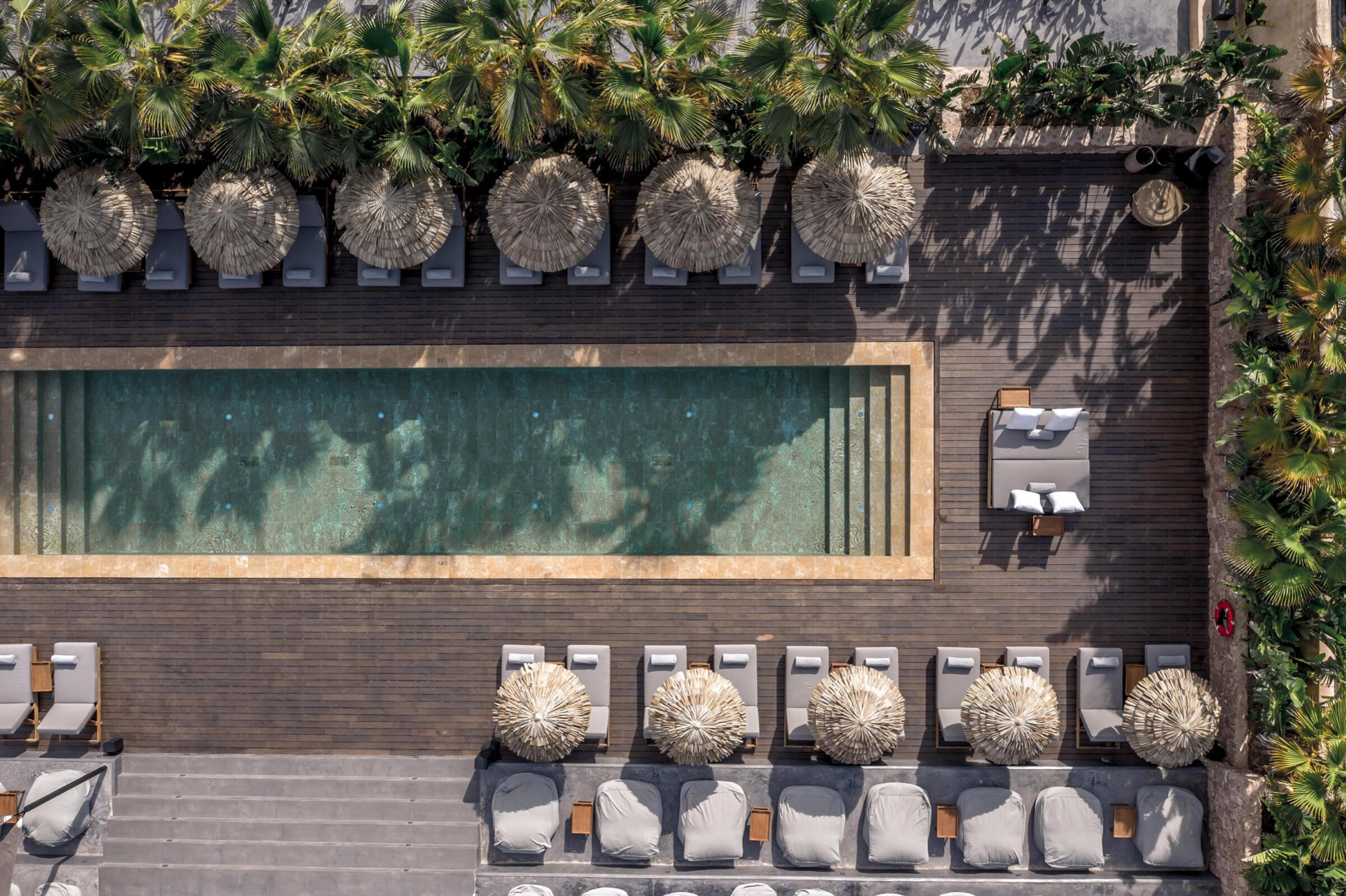Looking straight down on a long pool with a stone rim and dark green water, with white-cushioned loungers and thatched parasols along the timber decking and palms above.