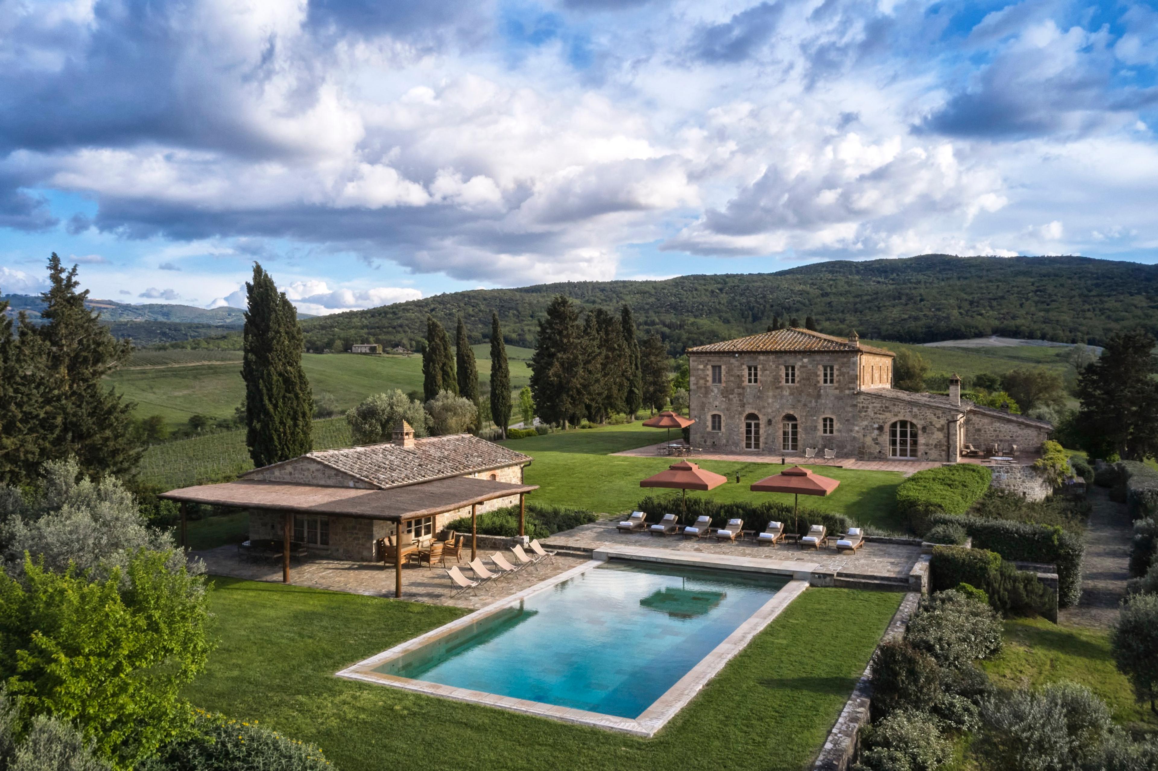 An aerial view of two stone buildings and a swimming pool with loungers and umbrellas, set among trees with hills and clouds behind.