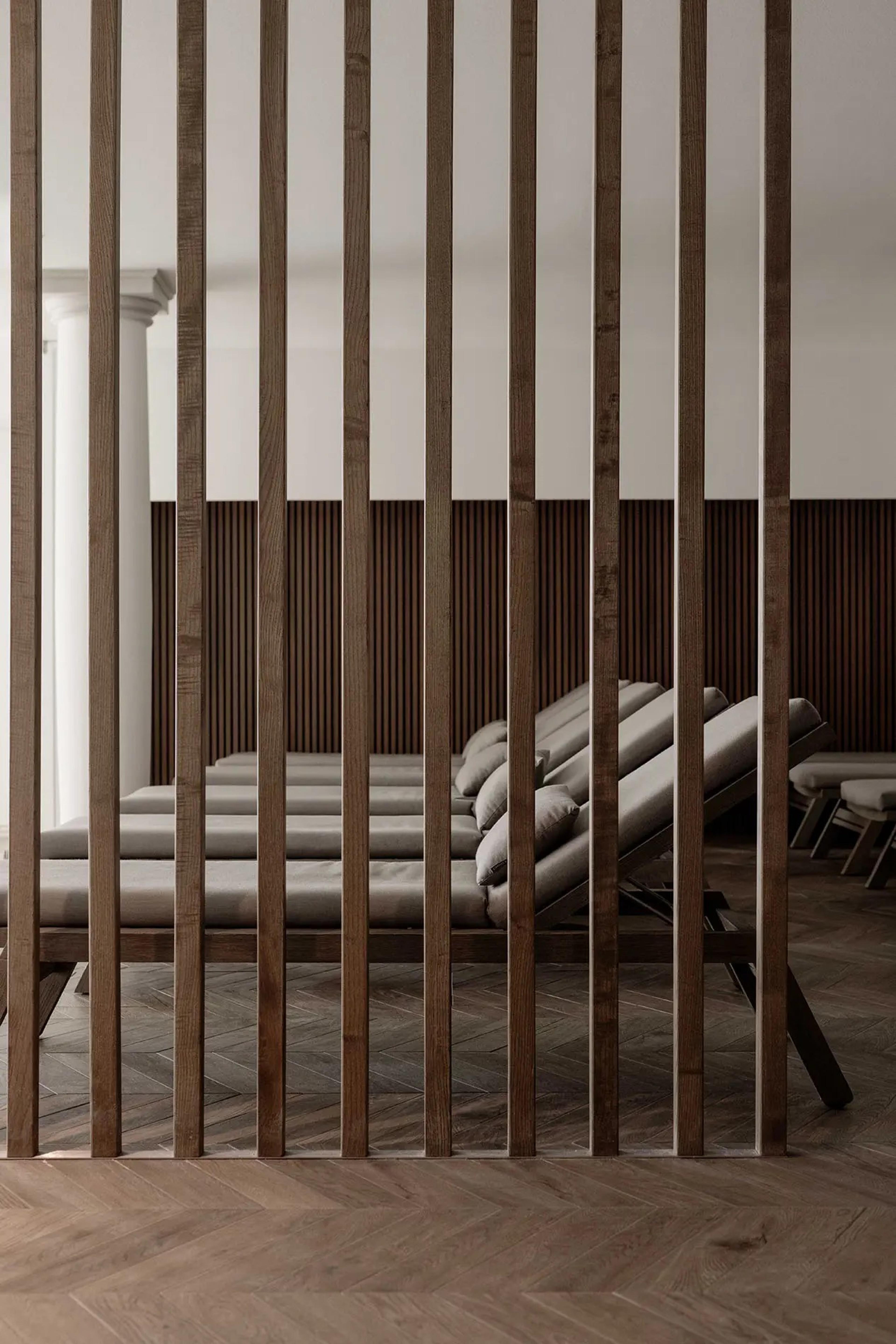 Wooden slats form a screen in front of a row of reclined loungers in a spa, with a stone column and herringbone floor behind.