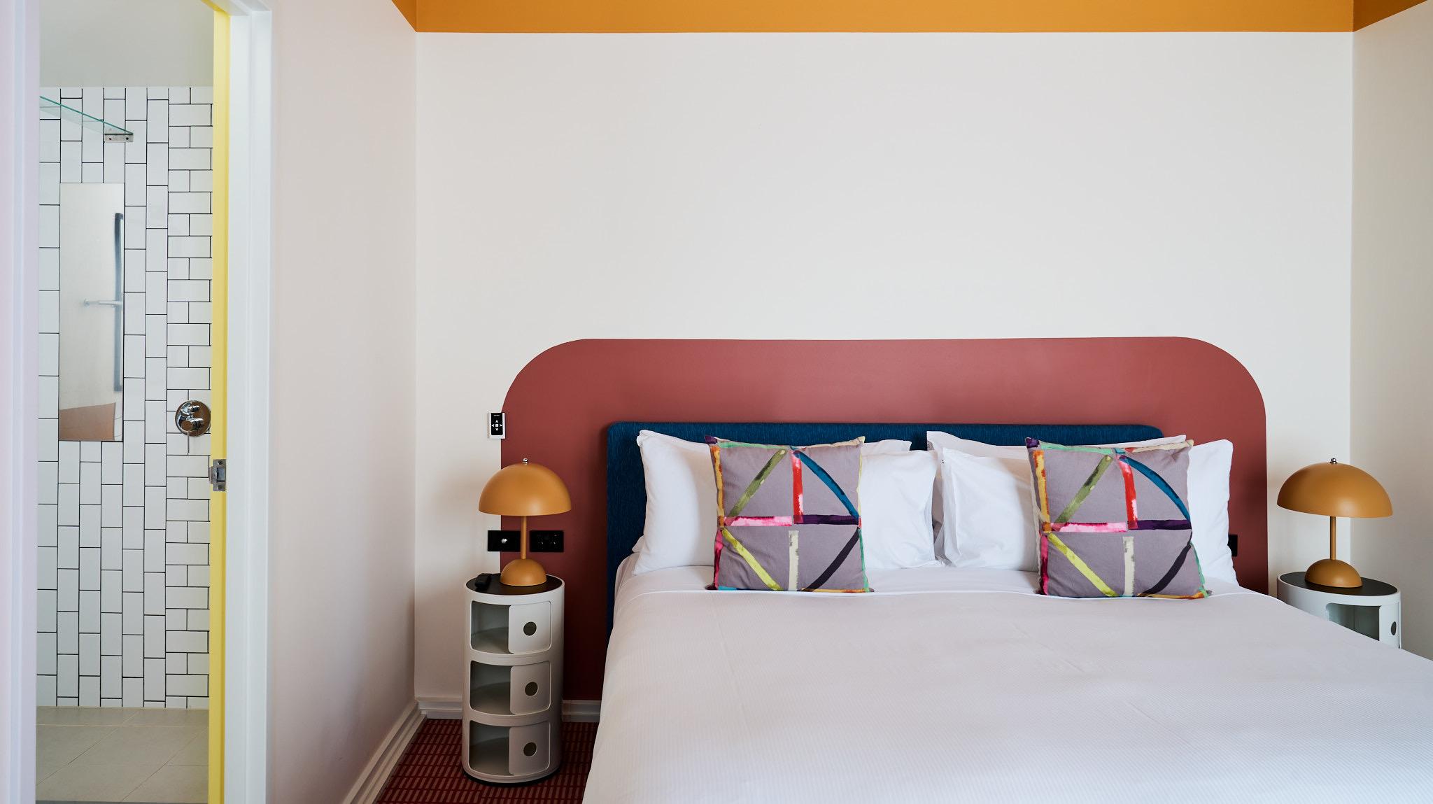 A bed with a maroon headboard and graphic cushions, gold bedside lamps and an open door to a tiled ensuite bathroom.