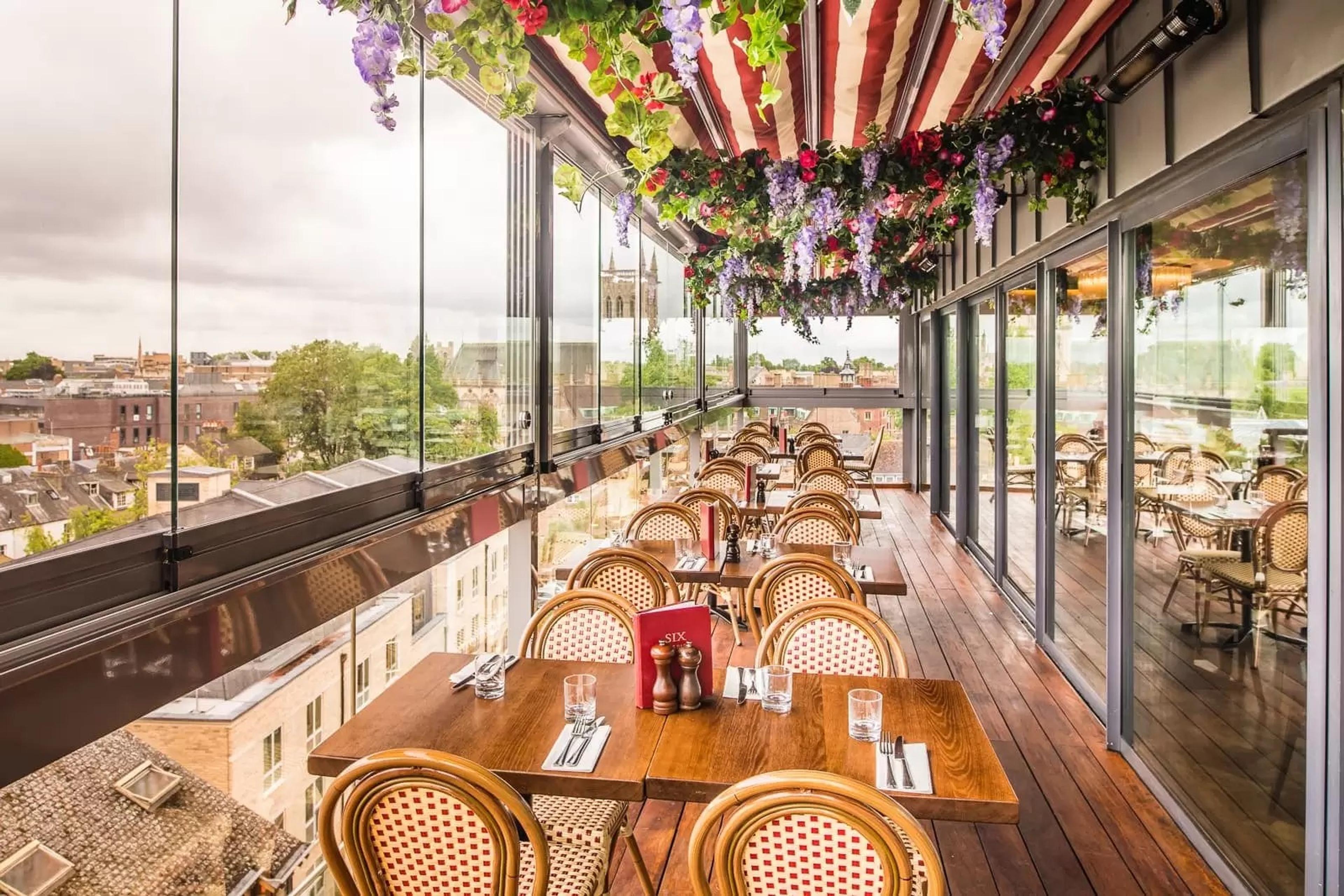 a restaurant balcony with tables set for lunch under a canopy hung with flowers, looking out over Cambridge rooftops and a church tower