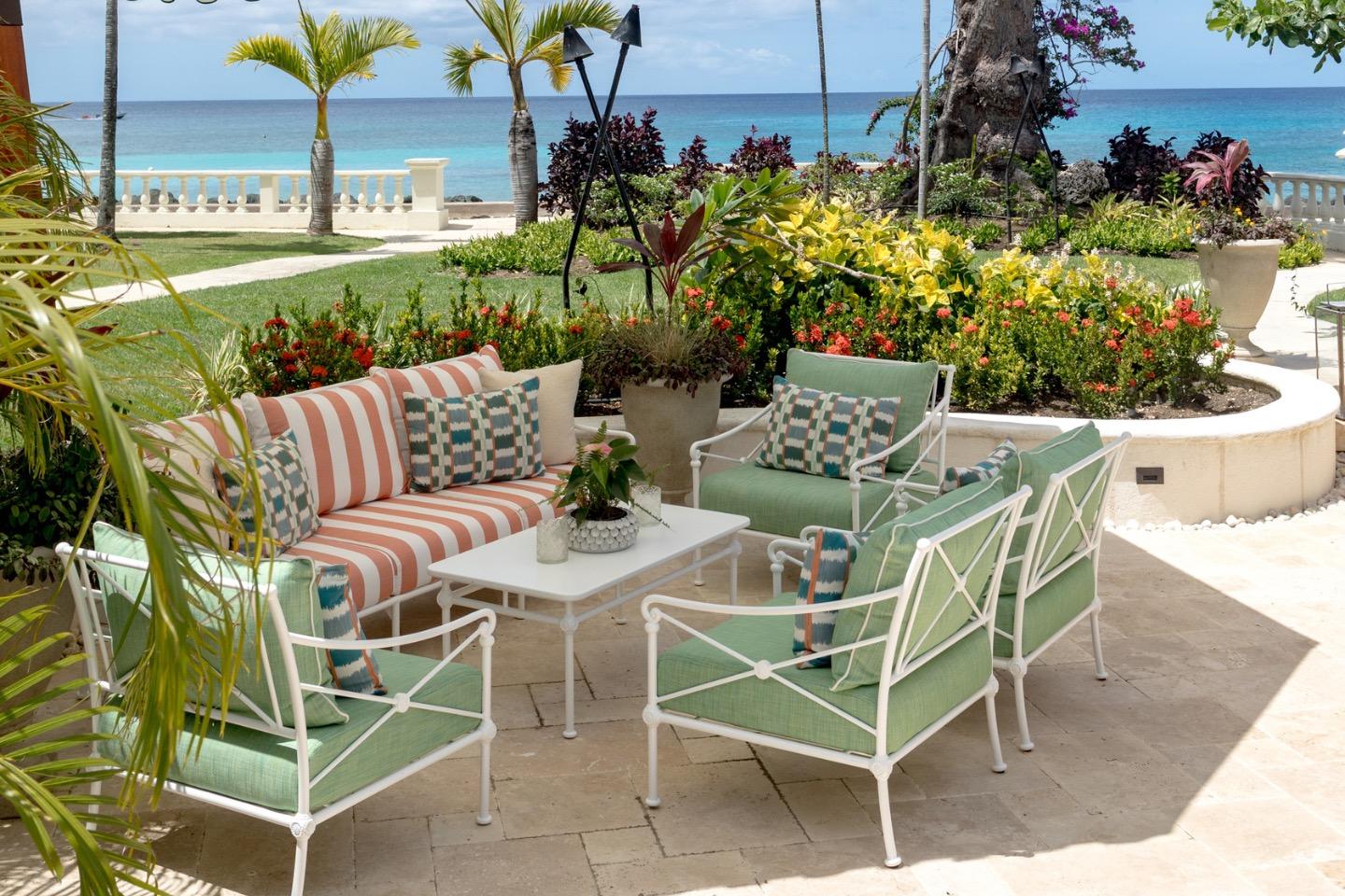 An outdoor seating area with striped and green cushioned chairs around a low table, set in a garden of red and yellow flowering shrubs with the sea beyond.